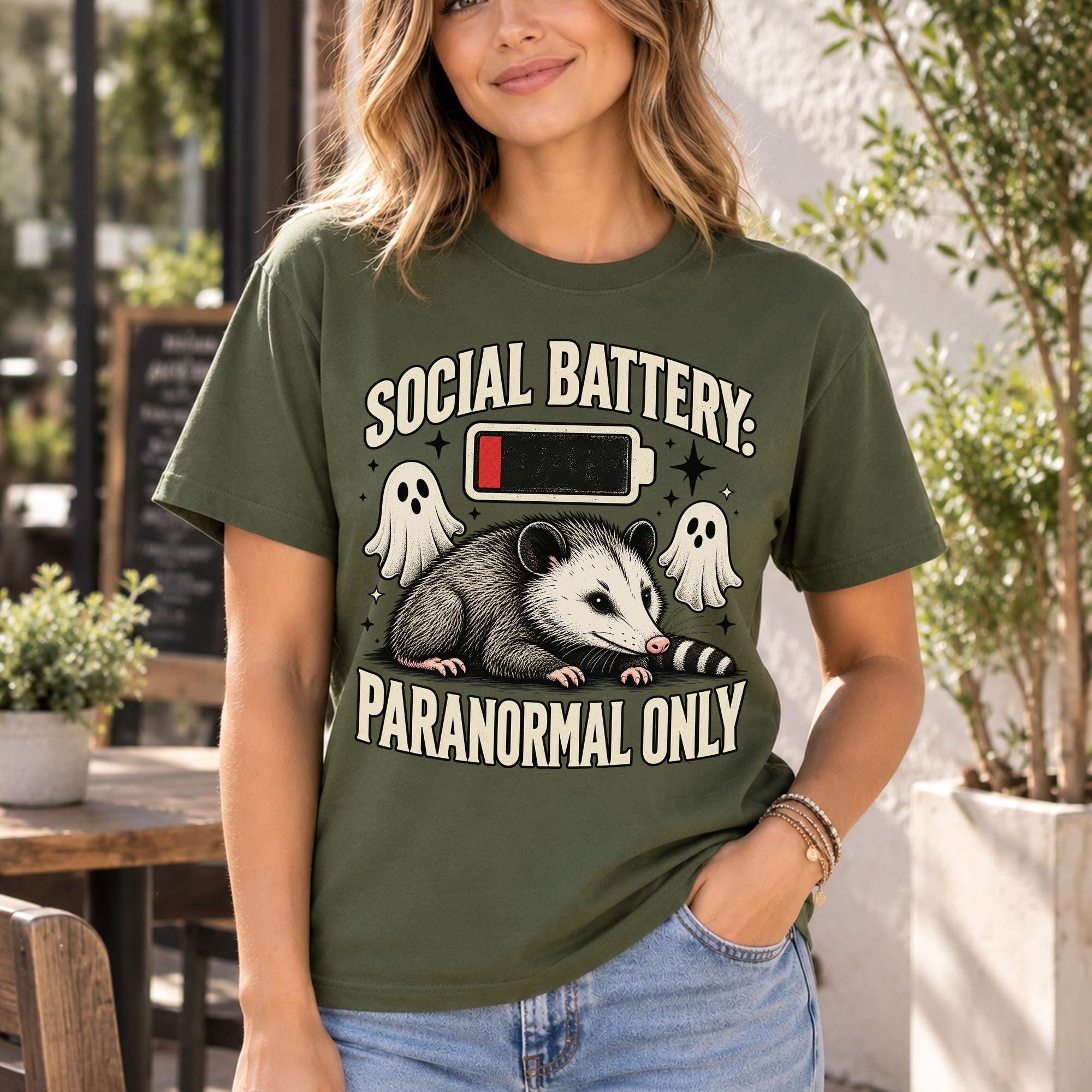 A woman wearing a green t-shirt with a graphic design that says "SOCIAL BATTERY: PARANORMAL ONLY" and features a cartoon opossum and ghost-like figures.