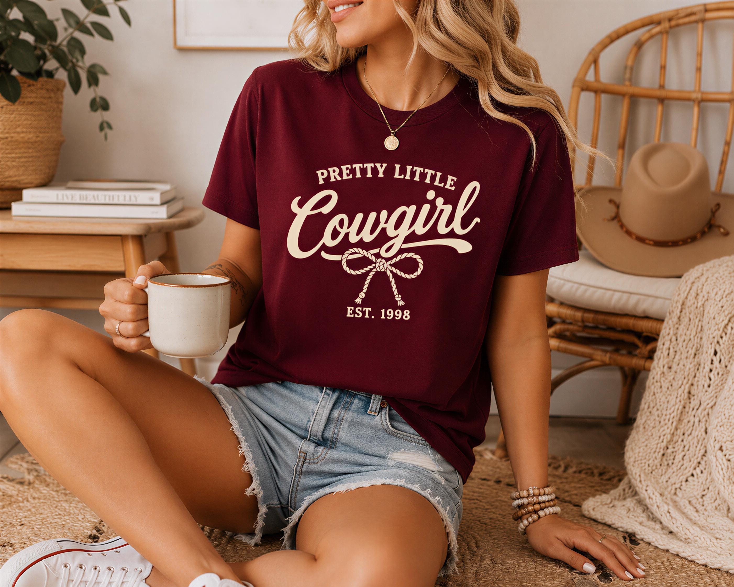 A woman wearing a maroon t-shirt with the text "PRETTY LITTLE COUGIRL" and denim shorts is sitting on the floor, holding a coffee mug and wearing a necklace.