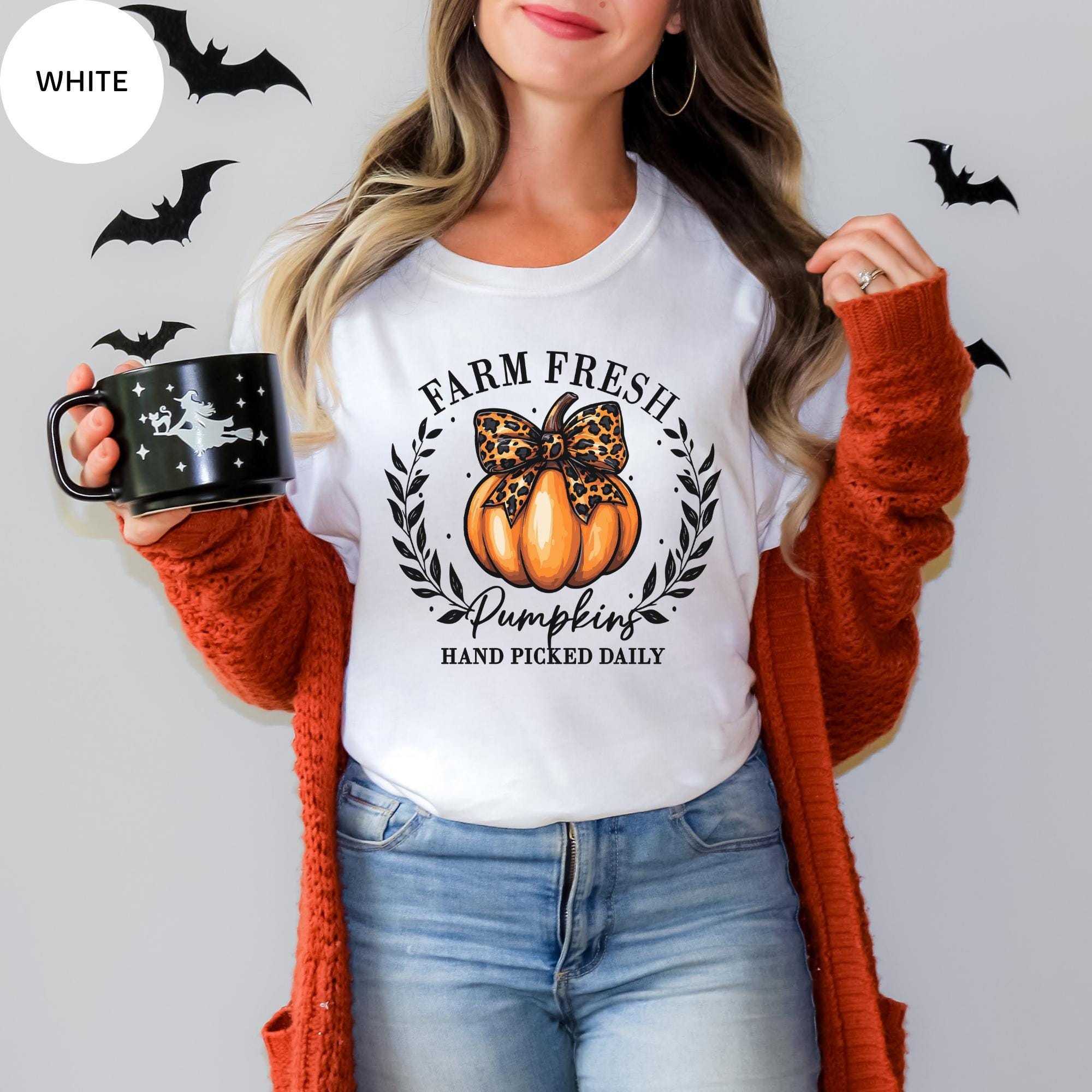 A woman wearing a white t-shirt with a pumpkin graphic and the text "Farm Fresh Pumpkins Hand Picked Daily" is holding a black mug with a pumpkin design. She is also wearing an orange cardigan and jeans.