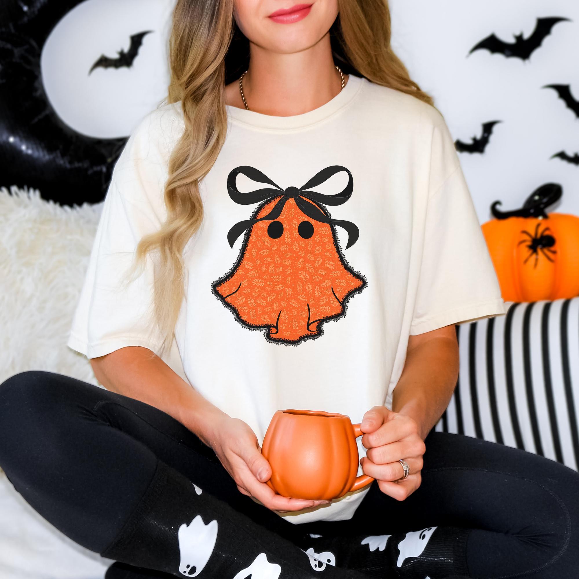 A woman wearing a white t-shirt with an orange ghost design and holding a pumpkin-shaped mug, sitting on a couch with Halloween decorations in the background.