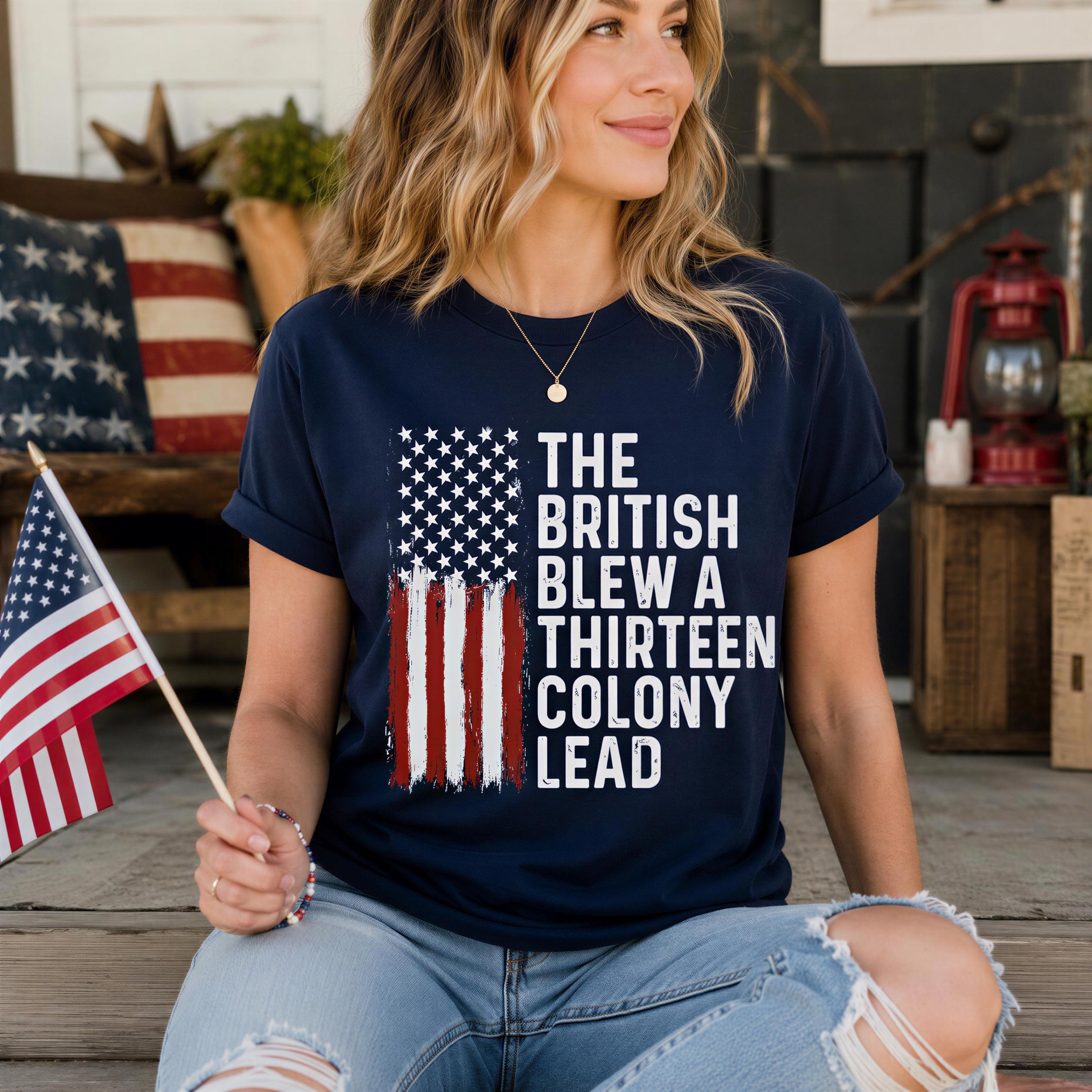 A woman wearing a navy blue t-shirt with the text "The British Blew a Thirteen Colony Lead" is sitting on a porch, holding an American flag.