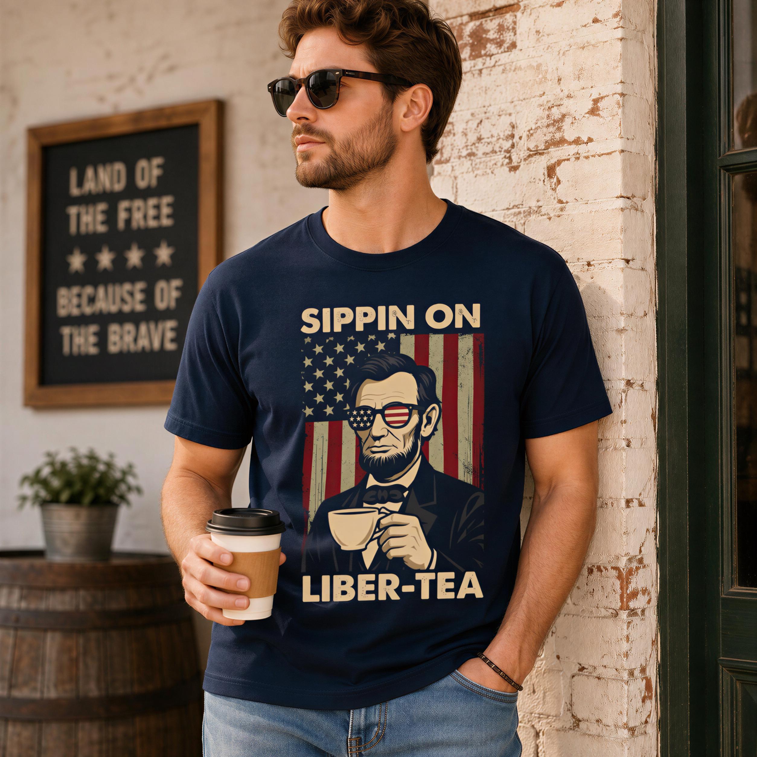 A man wearing a navy blue t-shirt with a graphic design of Abraham Lincoln and the text "SIPPIN ON LIBER-TEA" is holding a coffee cup and standing in front of a brick wall.