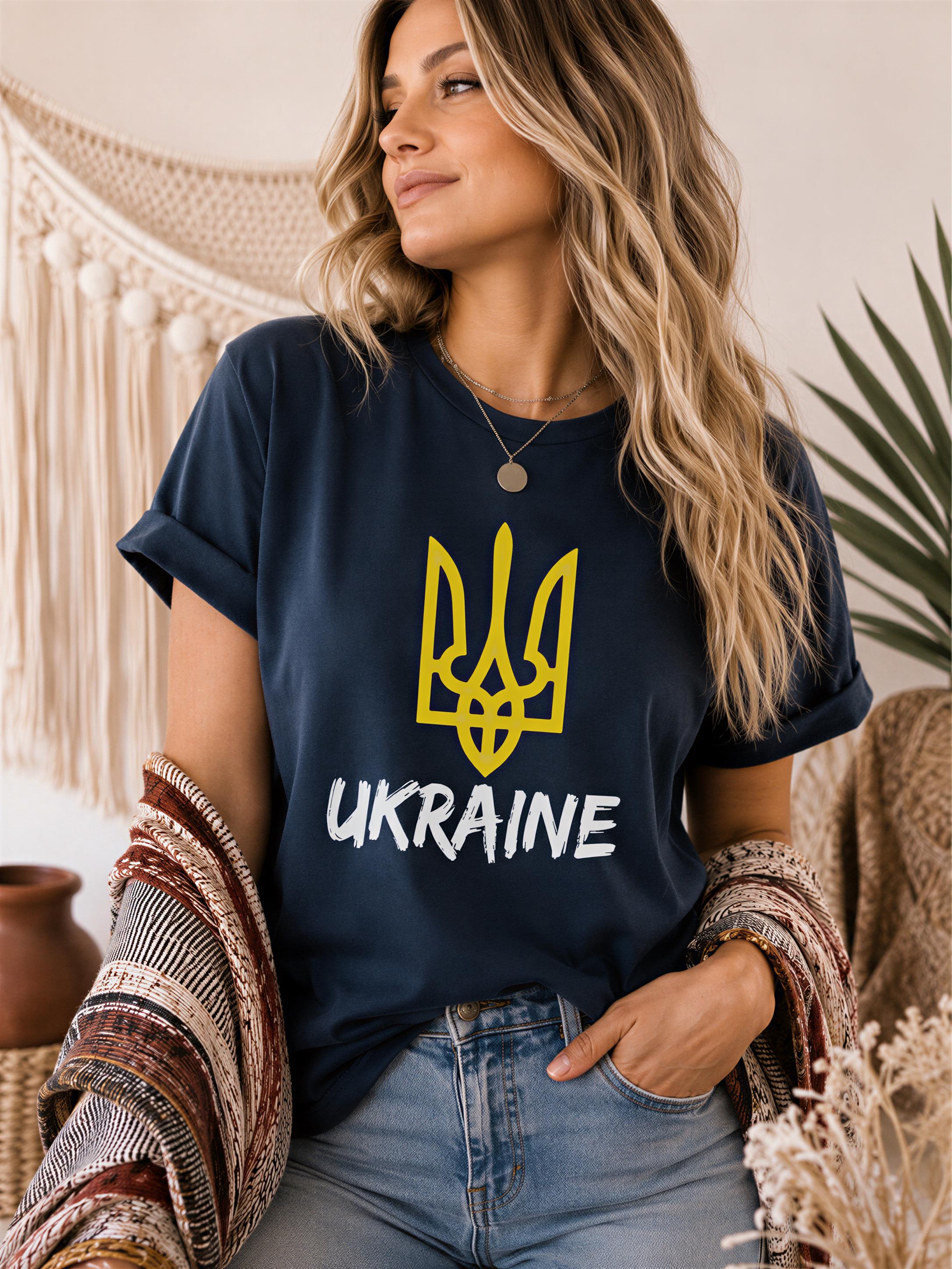 A woman wearing a navy blue t-shirt with the word "UKRAINE" printed on it, standing in a room with a woven wall hanging and a plant in the background.