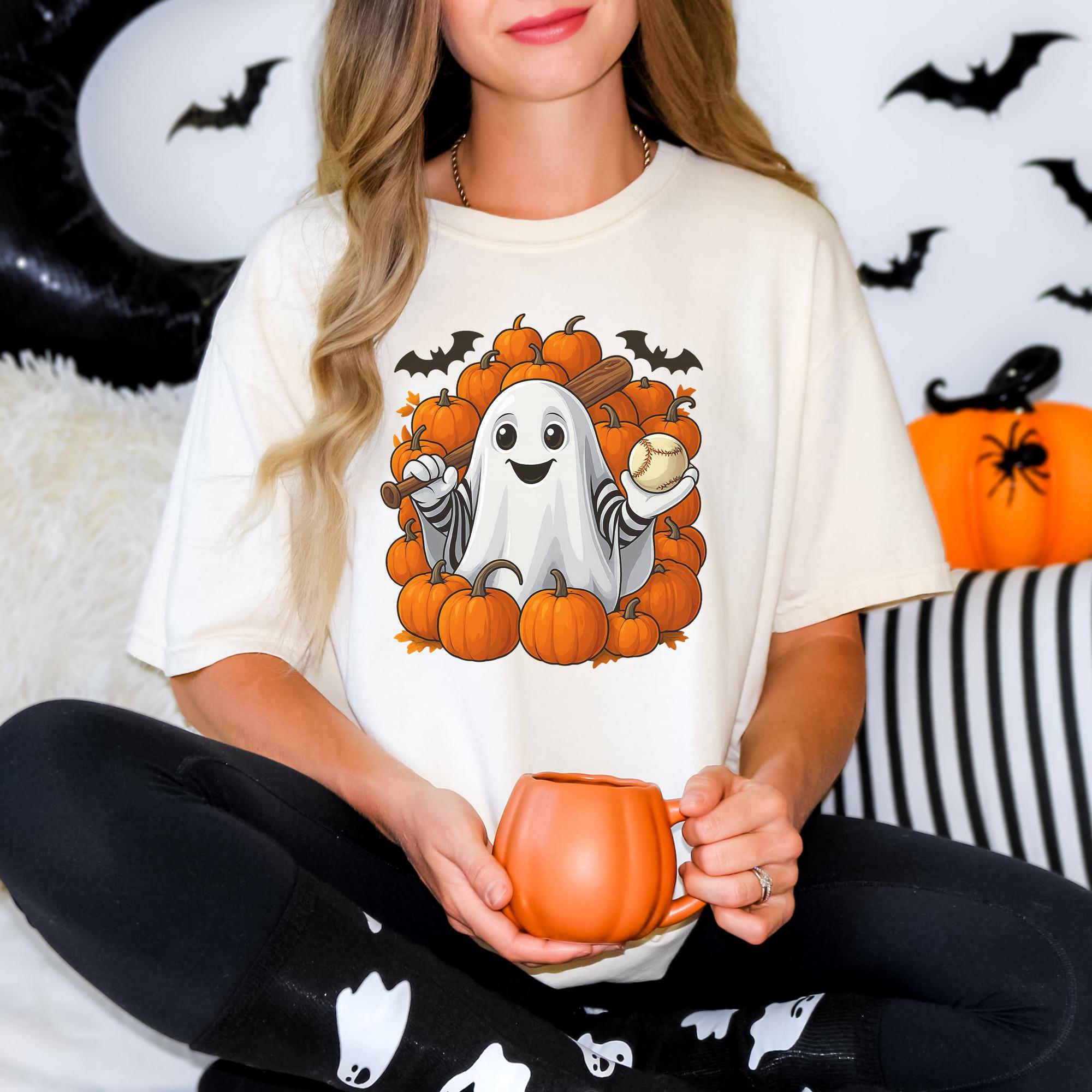 A woman wearing a white t-shirt with a graphic of a ghost holding a baseball bat and surrounded by pumpkins, sitting on a couch with a mug in her hand.