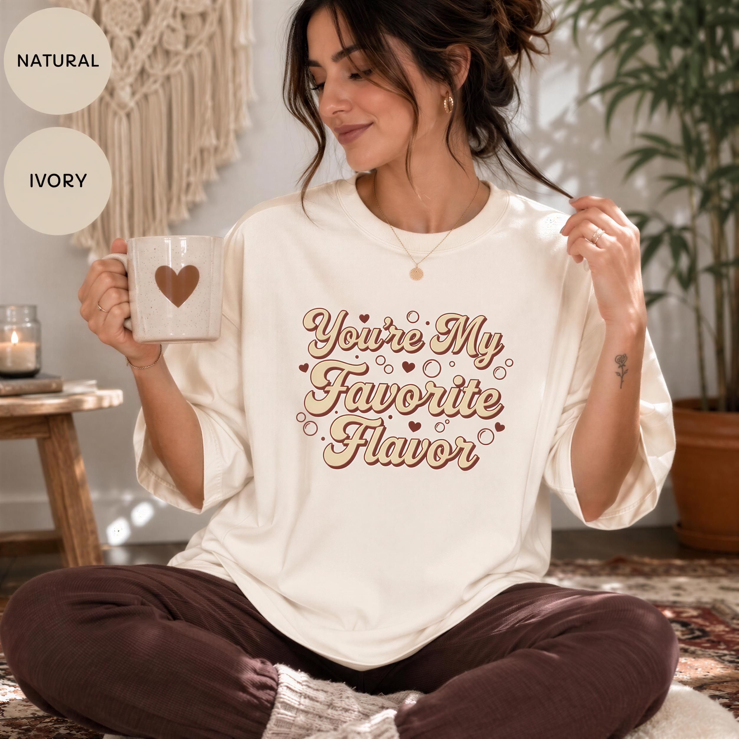 A woman sitting on the floor, holding a mug with a heart design, wearing a white t-shirt with a text design, and smiling.
