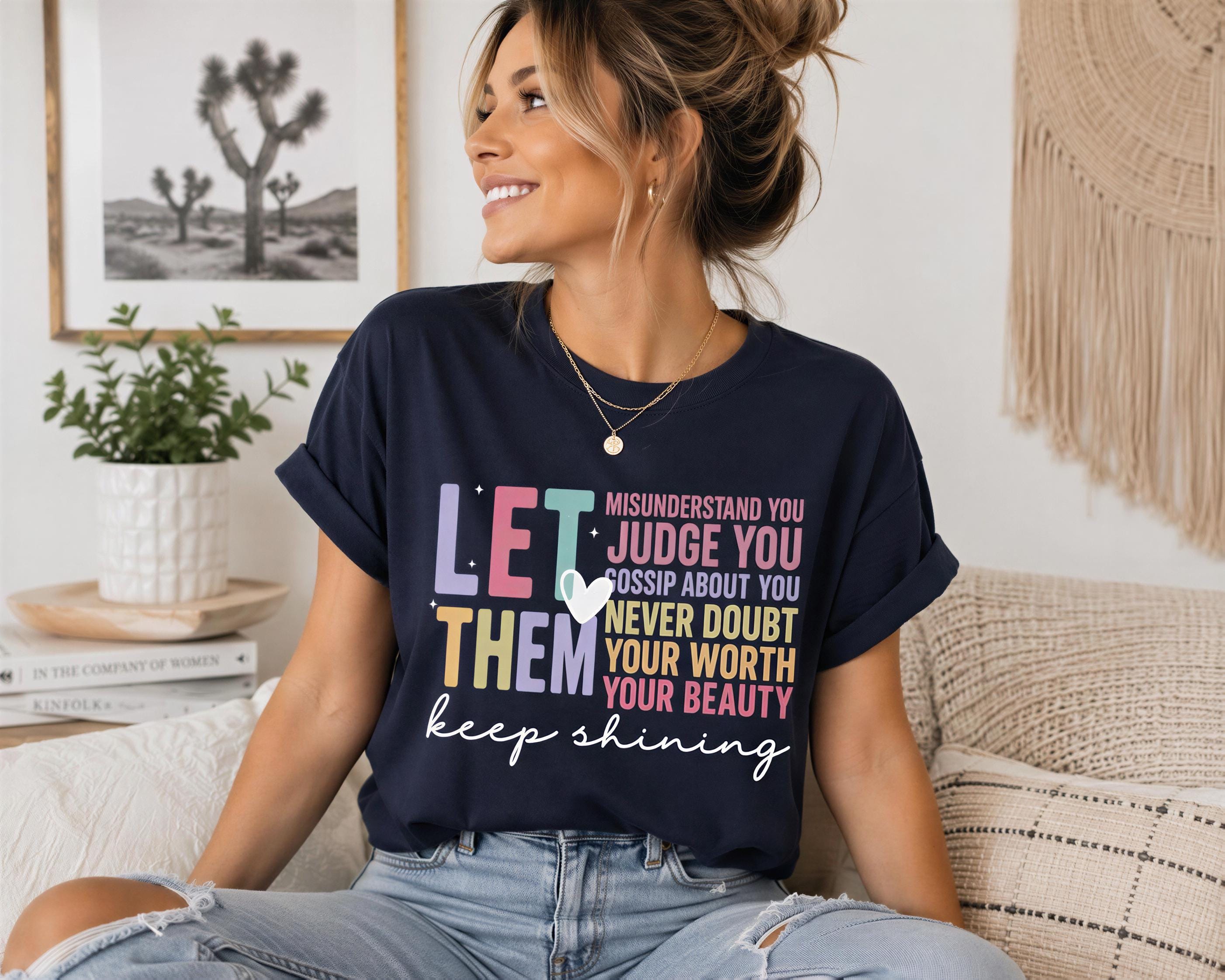 A woman wearing a black t-shirt with a colorful graphic design and text, sitting on a couch in a room with a potted plant and a framed picture on the wall.