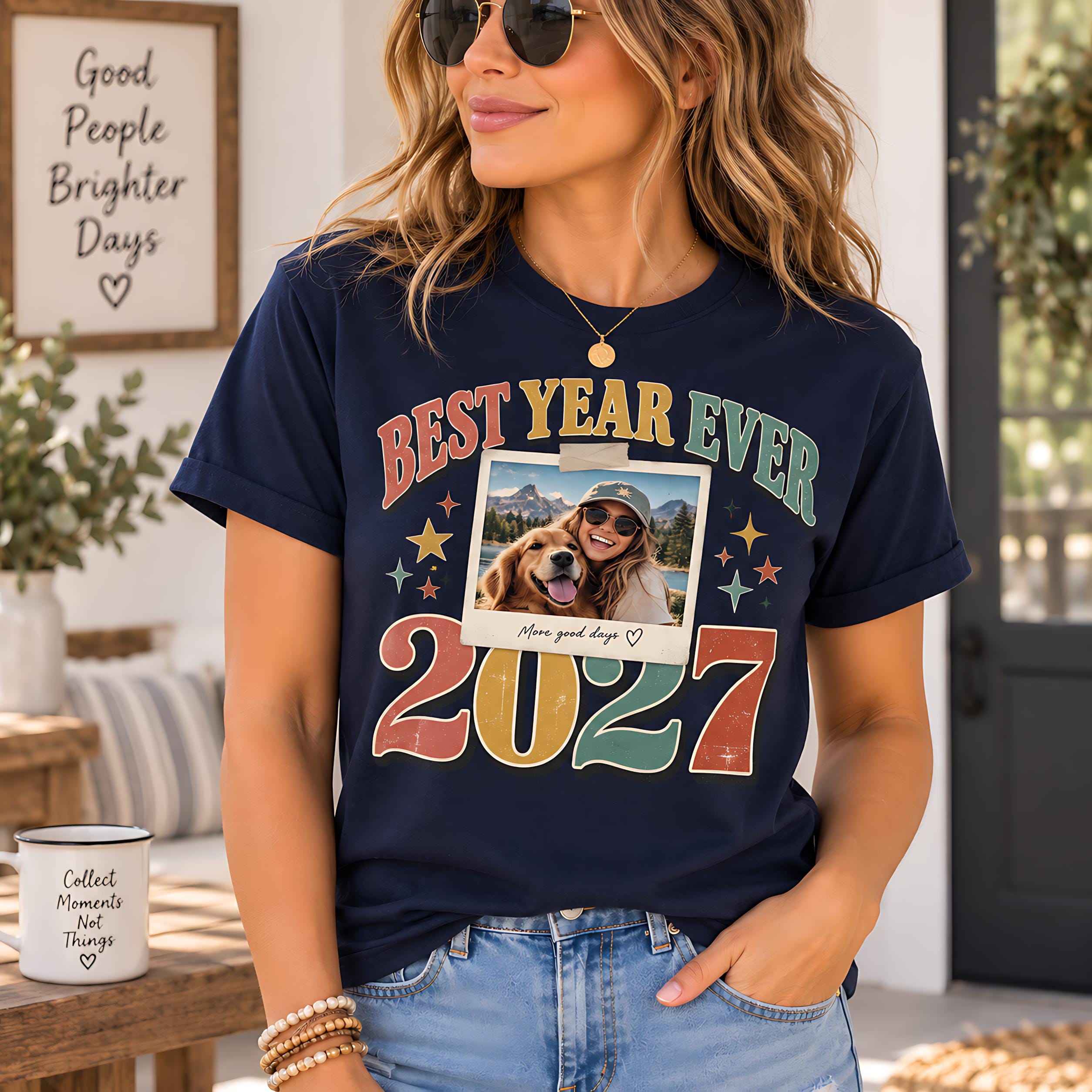 A woman wearing a navy blue t-shirt with a colorful graphic design and text, standing in front of a wooden table with a coffee cup on it.