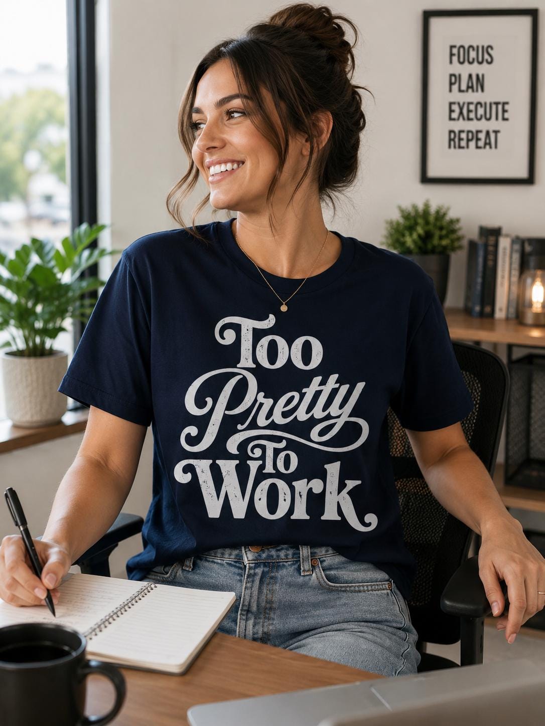 A woman wearing a navy blue t-shirt with the text "Too Pretty to Work" is sitting at a desk, smiling and holding a pen.