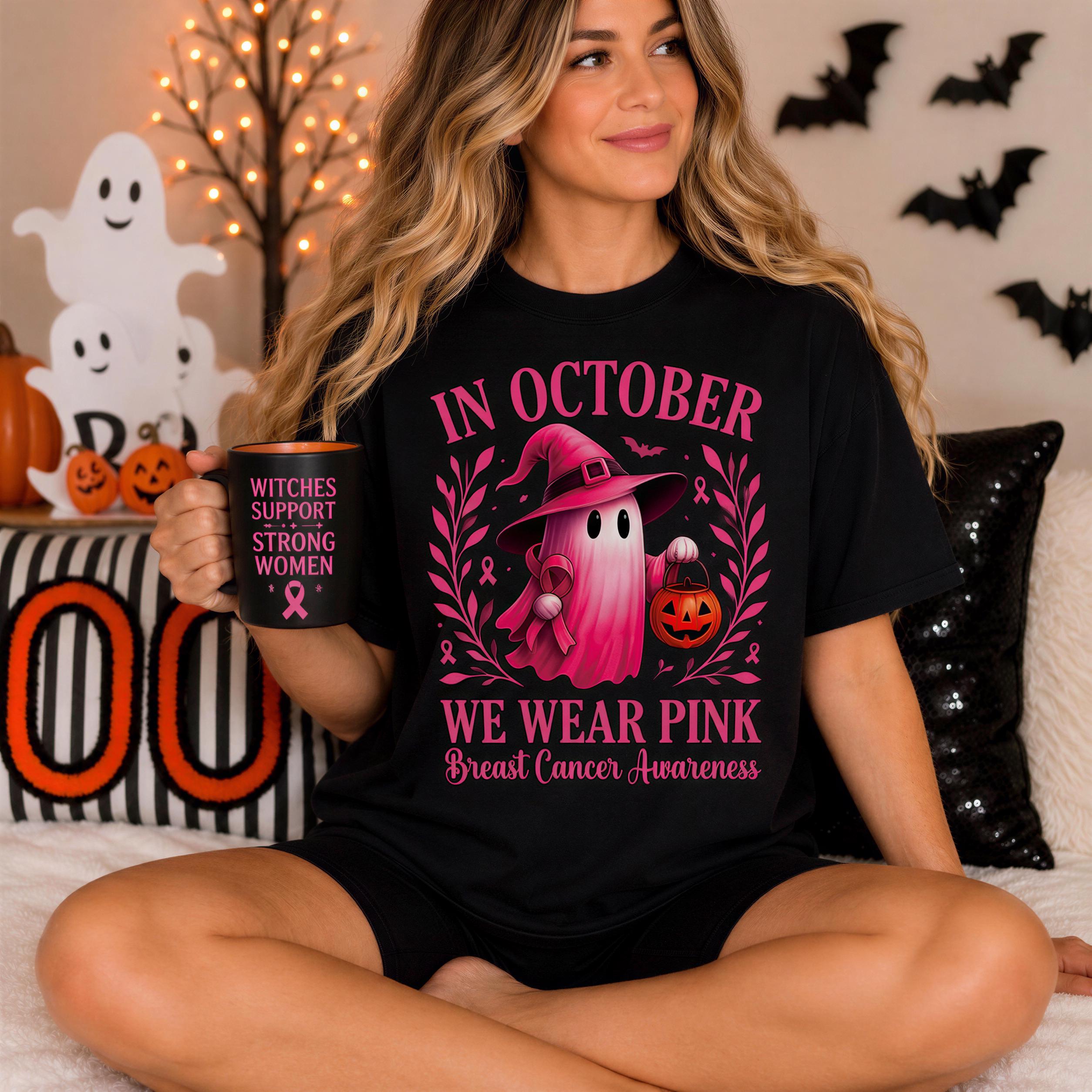 A woman wearing a black t-shirt with a graphic design of a pink ghost and the text "In October WE WEAR PINK Breast Cancer Awareness" is sitting on a bed. She is holding a mug with the text "Witches support strong women" printed on it.