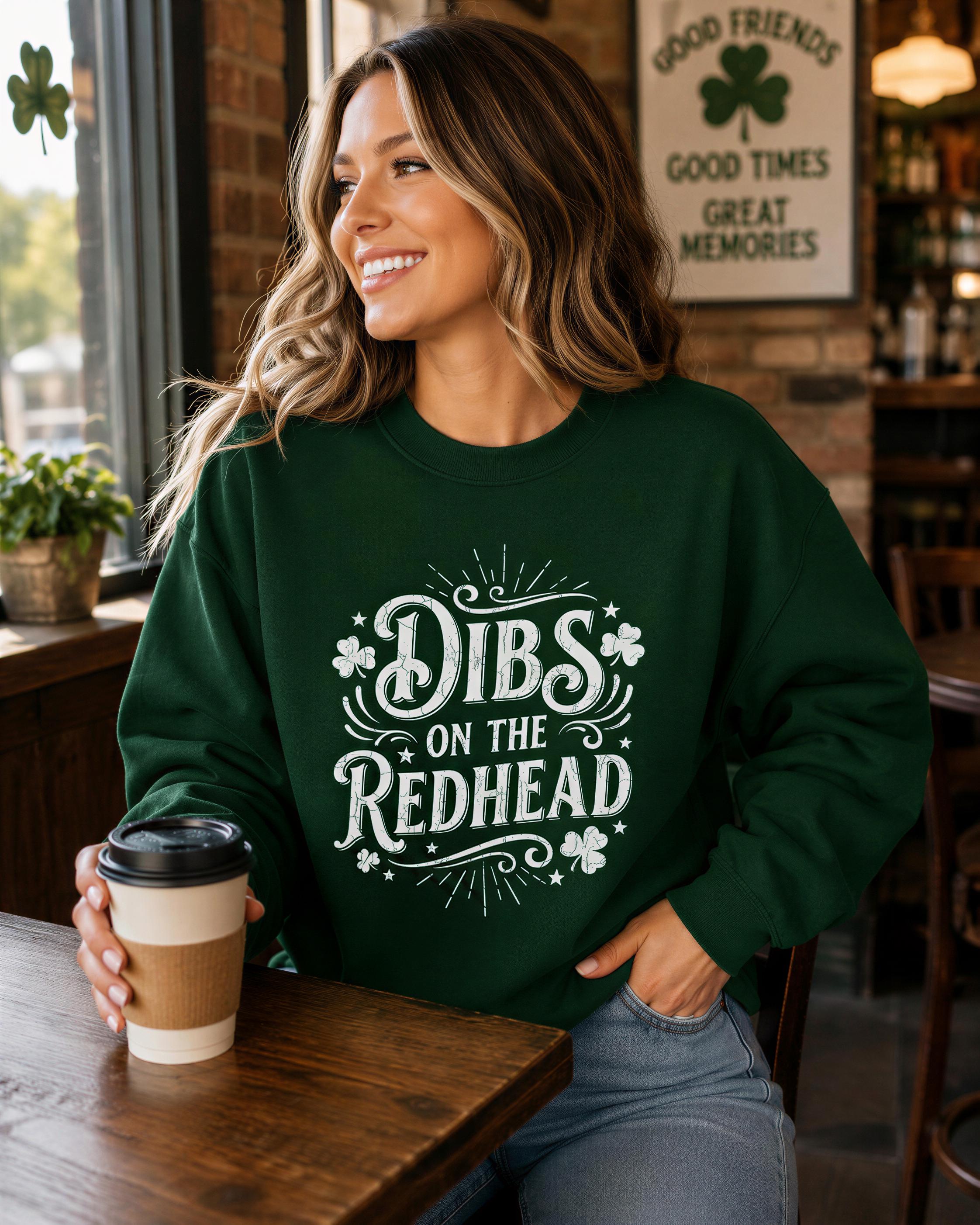 A woman wearing a green sweatshirt with the text "Dibs on the Redhead" is sitting at a table in a cafe, smiling and holding a coffee cup.