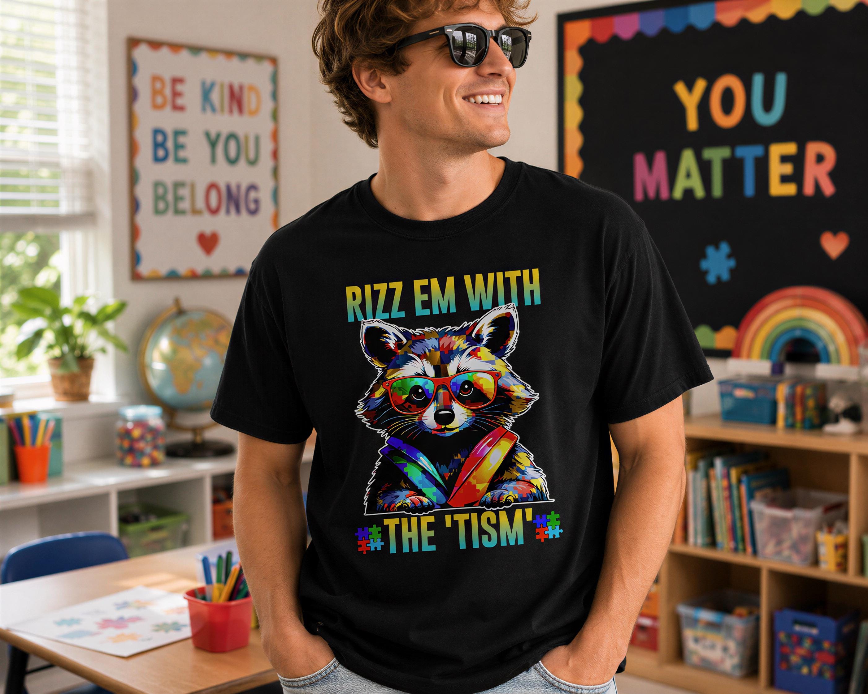 A man wearing a black t-shirt with a colorful raccoon graphic and text, standing in a classroom with a rainbow-colored wall behind him.