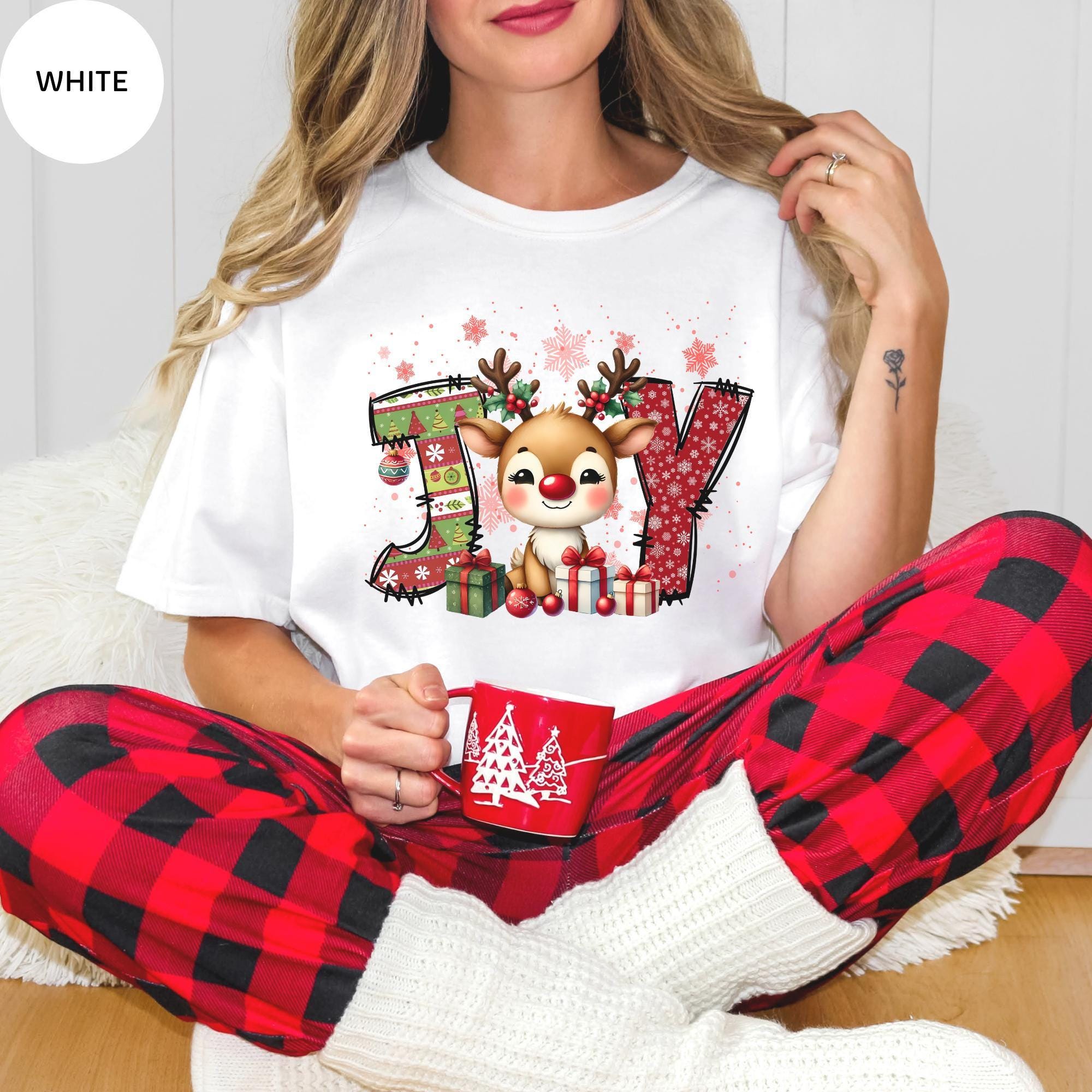 A woman wearing a white t-shirt with a Christmas-themed graphic and red plaid pajama bottoms, holding a red mug.