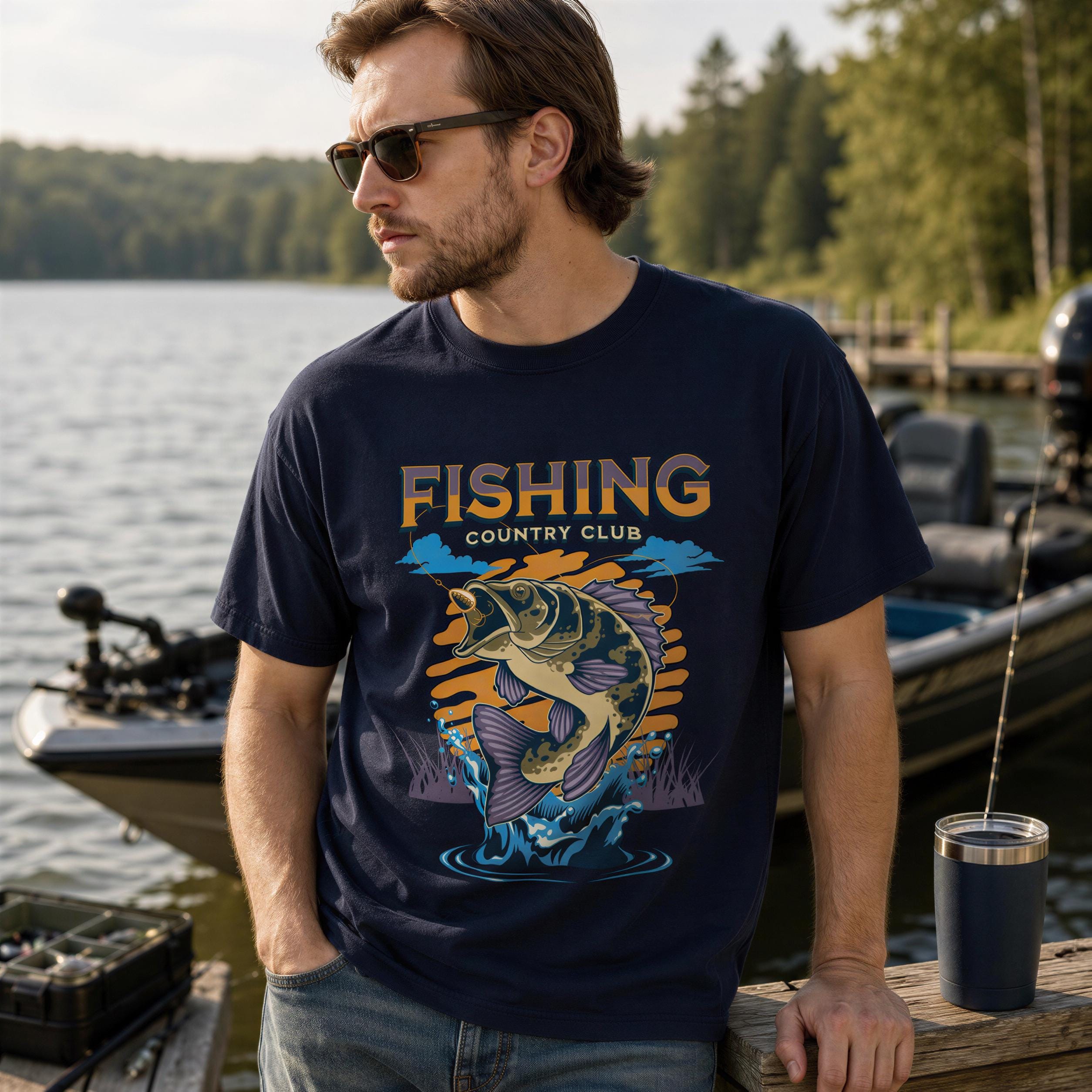 A man wearing a blue t-shirt with a fishing graphic stands near a lake, with a boat and trees in the background.