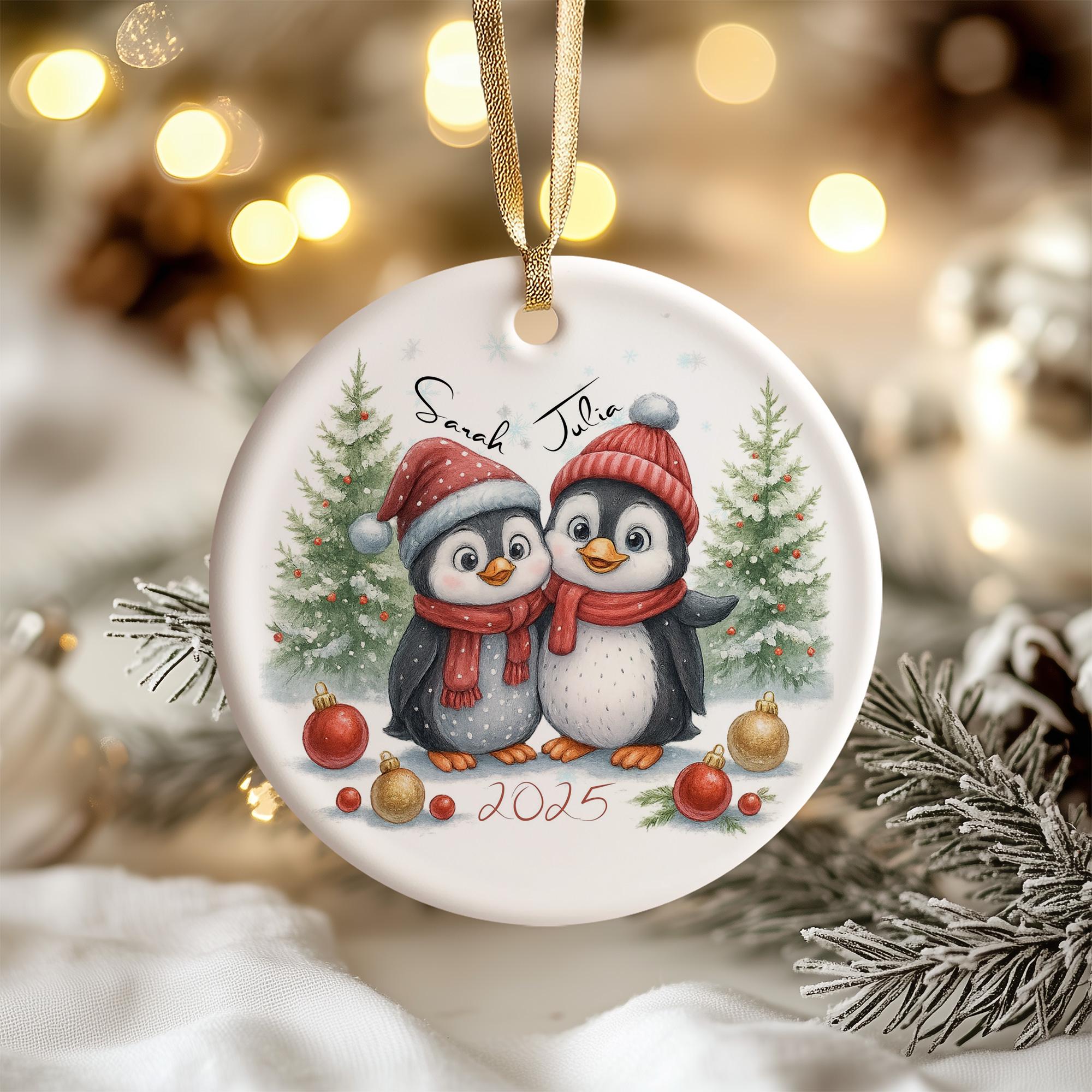 A white ornament with a picture of two penguins wearing red hats and scarves, surrounded by Christmas decorations such as ornaments and pine branches.