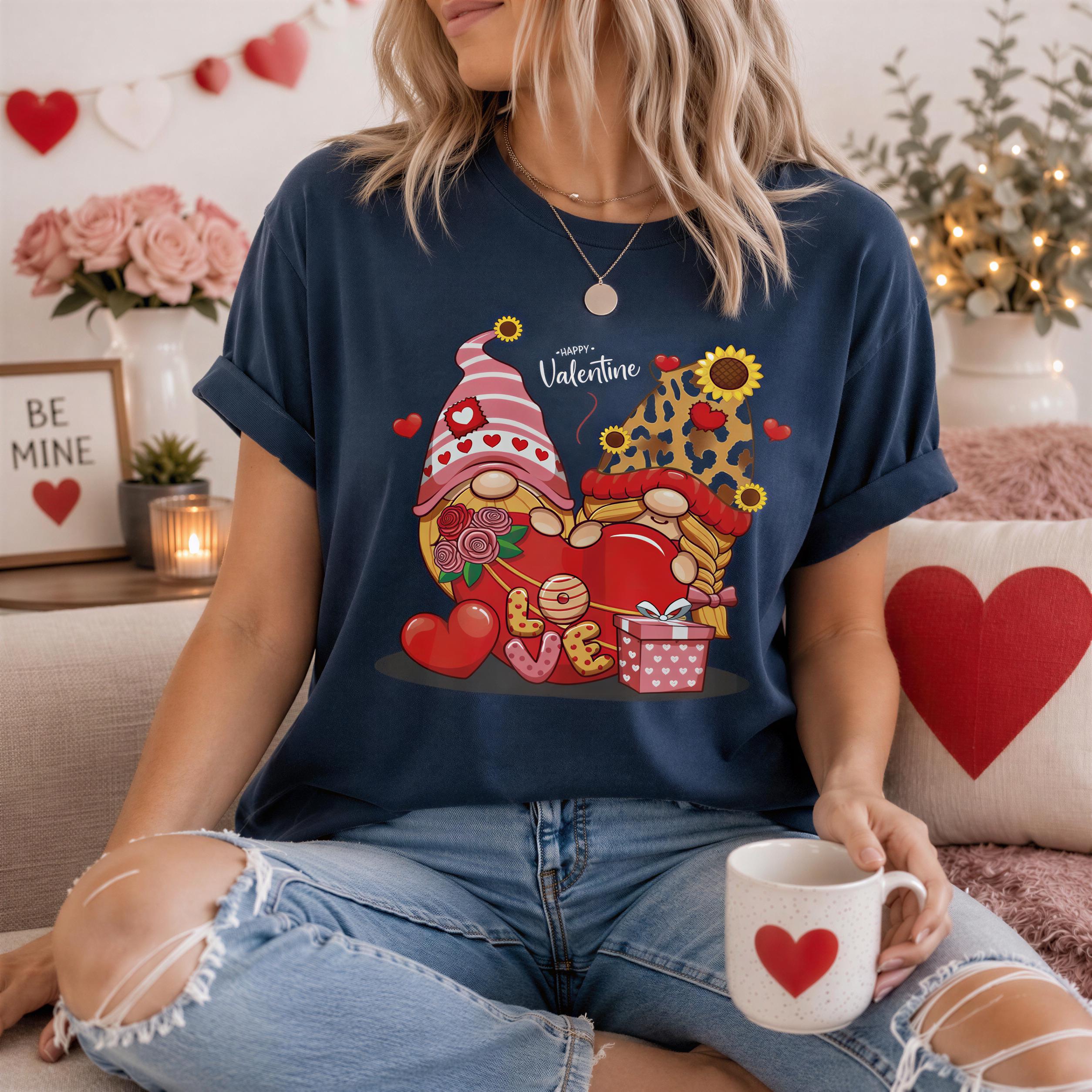 A woman wearing a blue t-shirt with a graphic of two gnomes holding hearts and flowers, sitting on a couch with a mug in her hand.