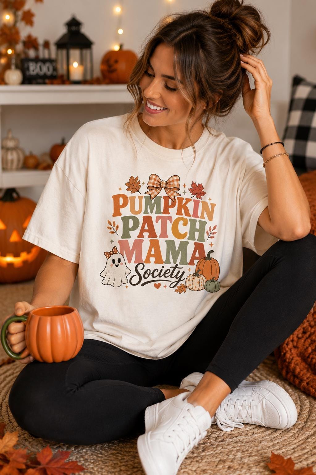 A woman sitting on the floor, wearing a white t-shirt with a pumpkin patch design, holding a pumpkin-shaped mug, and surrounded by fall decorations.