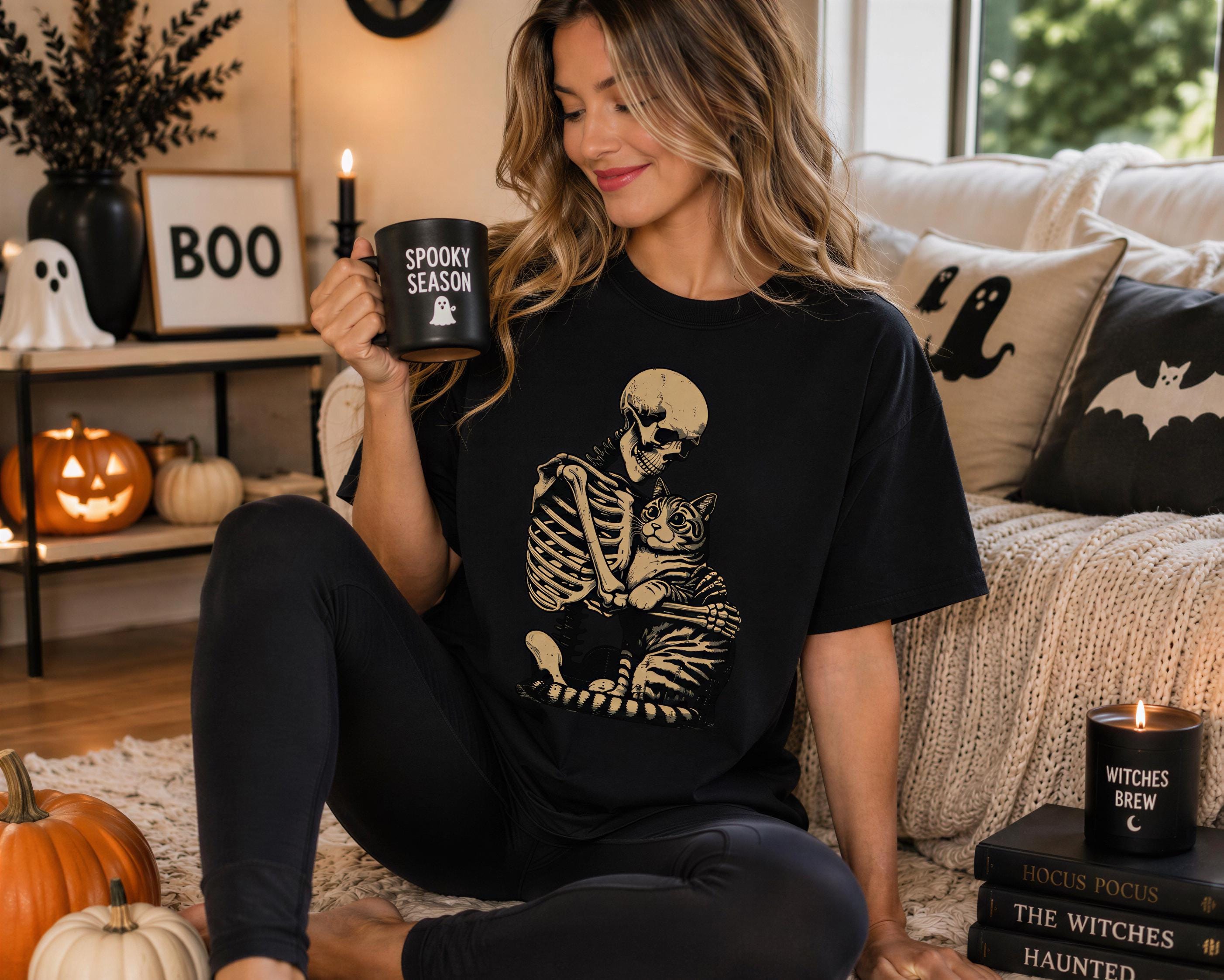 A woman in a black t-shirt with a skeleton graphic is sitting on the floor, holding a mug with the text "SPOOKY SEASON" and a cat graphic. Halloween decorations are visible in the background, including a skeleton decoration, a pumpkin, and a candle labeled "WITCHES BREW".