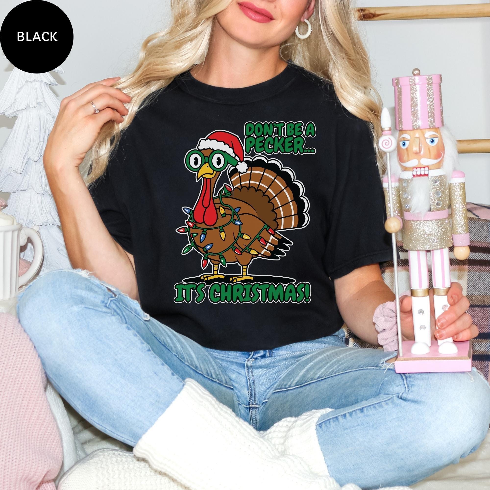 A woman wearing a black t-shirt with a graphic of a turkey wearing a Santa hat and the text "Don't be a PECKER! It's Christmas!" is sitting on a couch, holding a nutcracker.