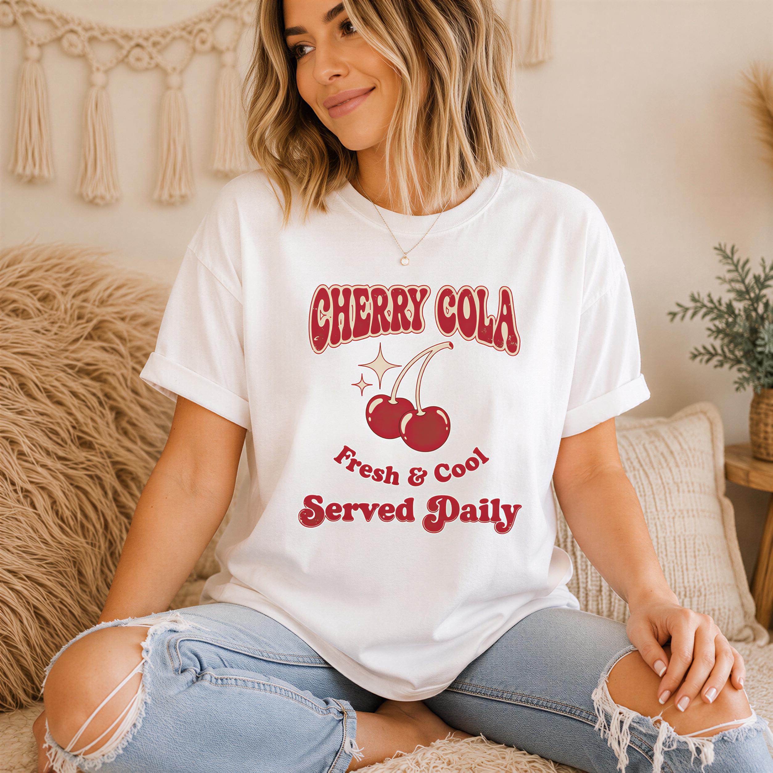 A woman wearing a white t-shirt with a graphic design of cherries and the text "CHERRY COLA" is sitting on a couch.