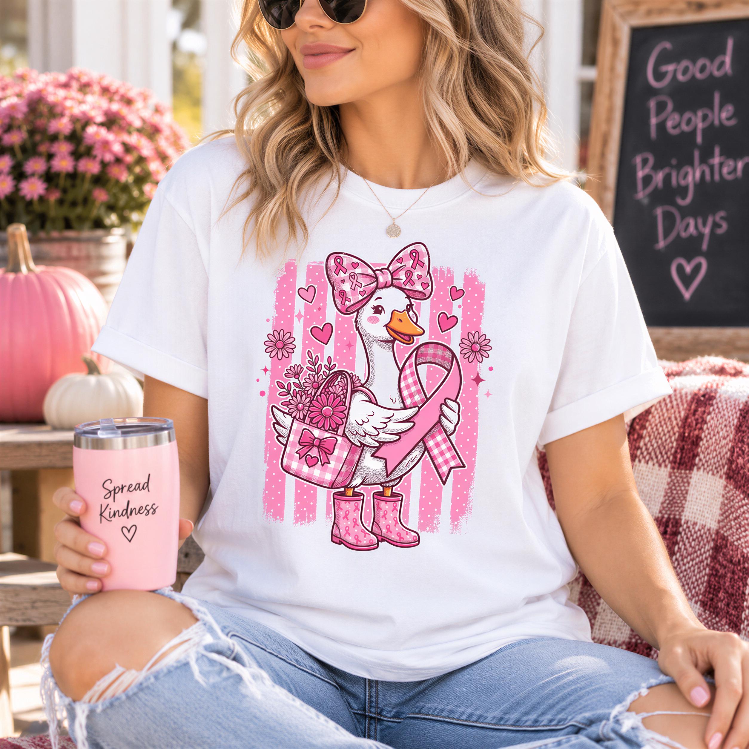 A woman wearing a white t-shirt with a pink graphic of a duck holding flowers and a pink ribbon, sitting on a bench and holding a pink cup with the text "Spread Kindness" printed on it.
