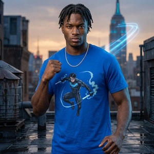 Peut inclure: Un t-shirt bleu vif à manches courtes avec une impression graphique d'un personnage de bande dessinée en costume noir avec un emblème d'oiseau bleu, tenant des bâtons lumineux. Le personnage est entouré de traînées d'énergie bleu clair.