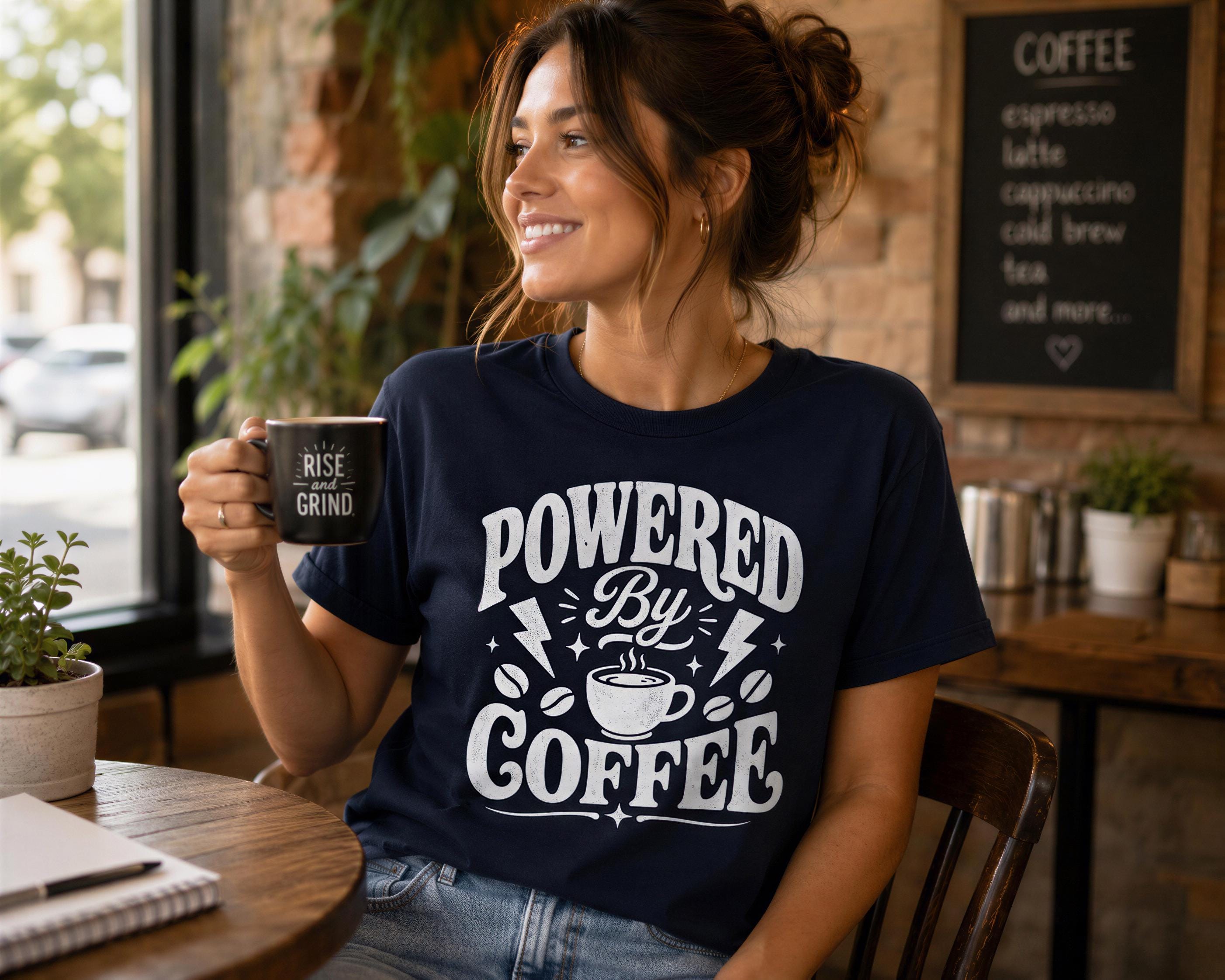 A woman wearing a navy blue t-shirt with the text "POWERED BY COFFEE" is sitting at a table in a coffee shop, holding a coffee mug and smiling.