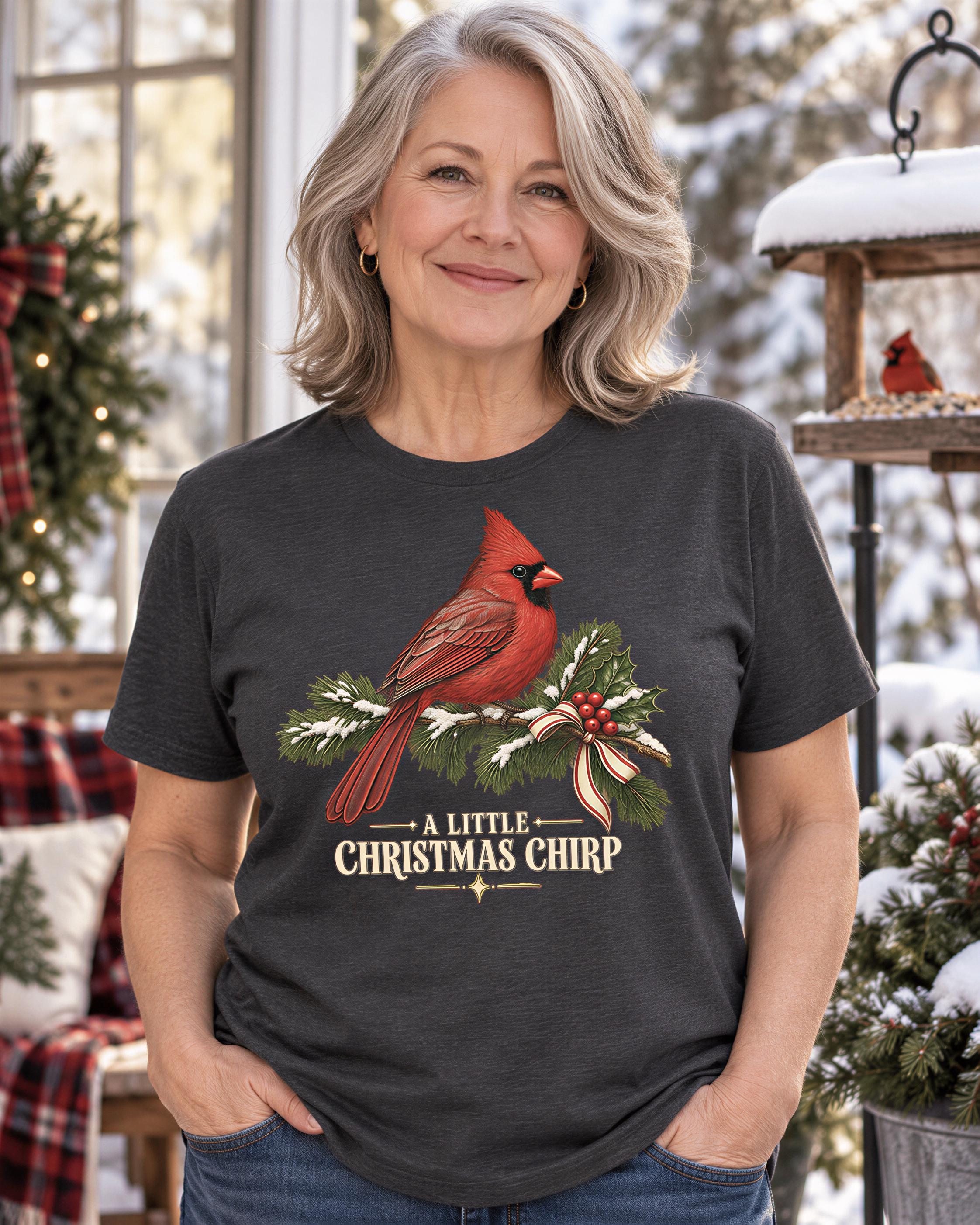 A woman wearing a gray t-shirt with a red cardinal bird graphic and the text "A little Christmas chip" is standing in front of a snowy background with Christmas decorations.