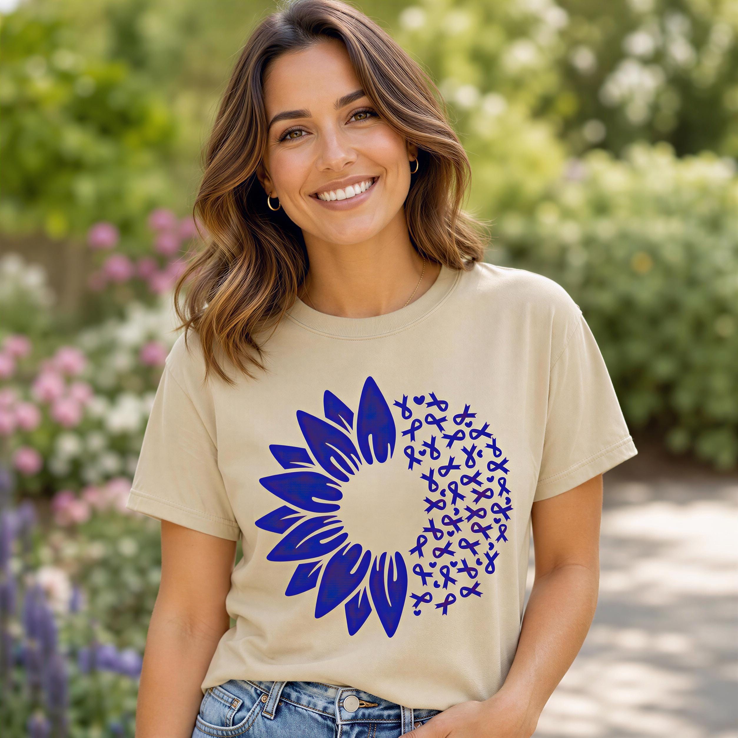 A smiling woman wearing a beige t-shirt with a blue sunflower design and blue ribbon pattern.