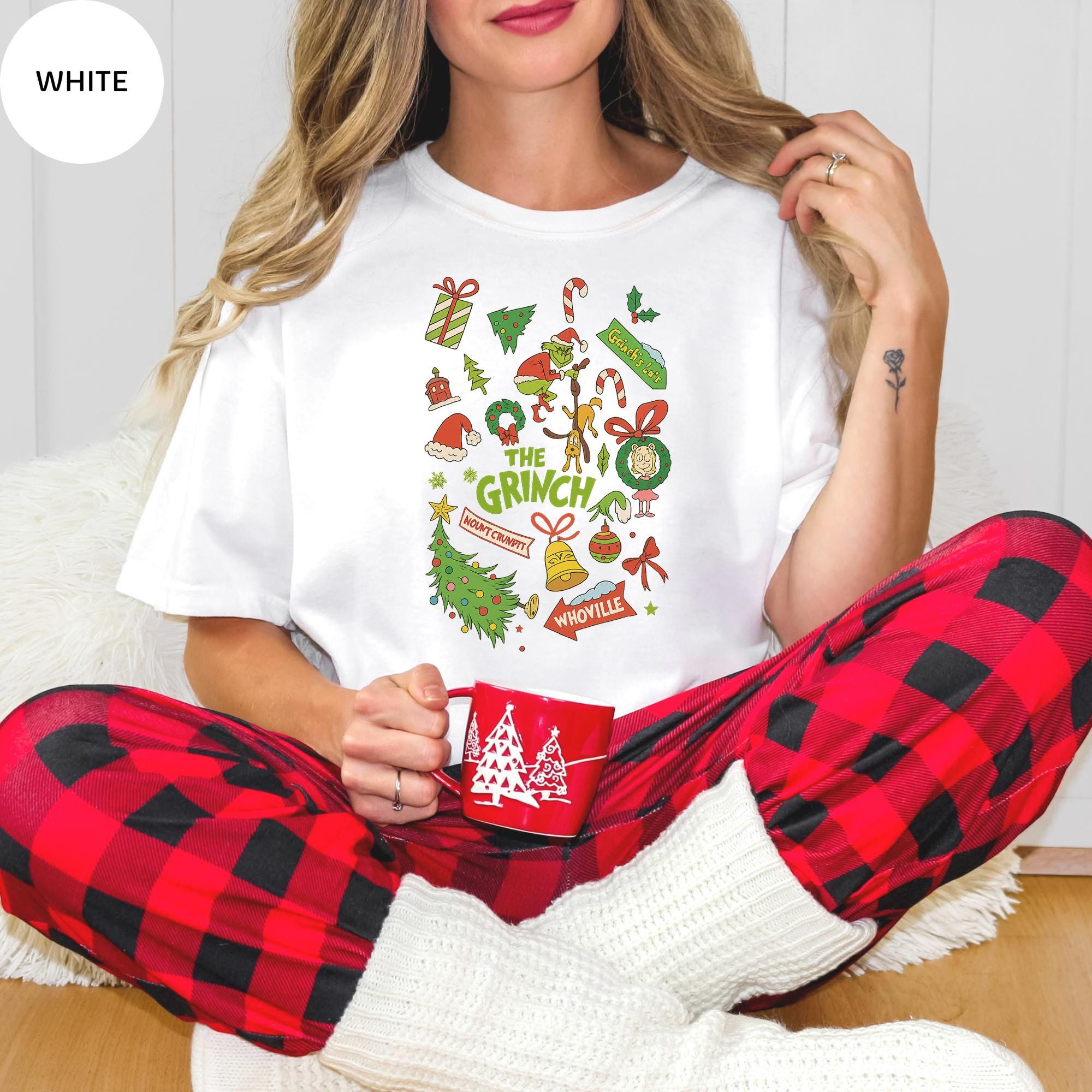 A woman wearing a white t-shirt with a graphic design and red plaid pajama bottoms, holding a red mug, and sitting on the floor.