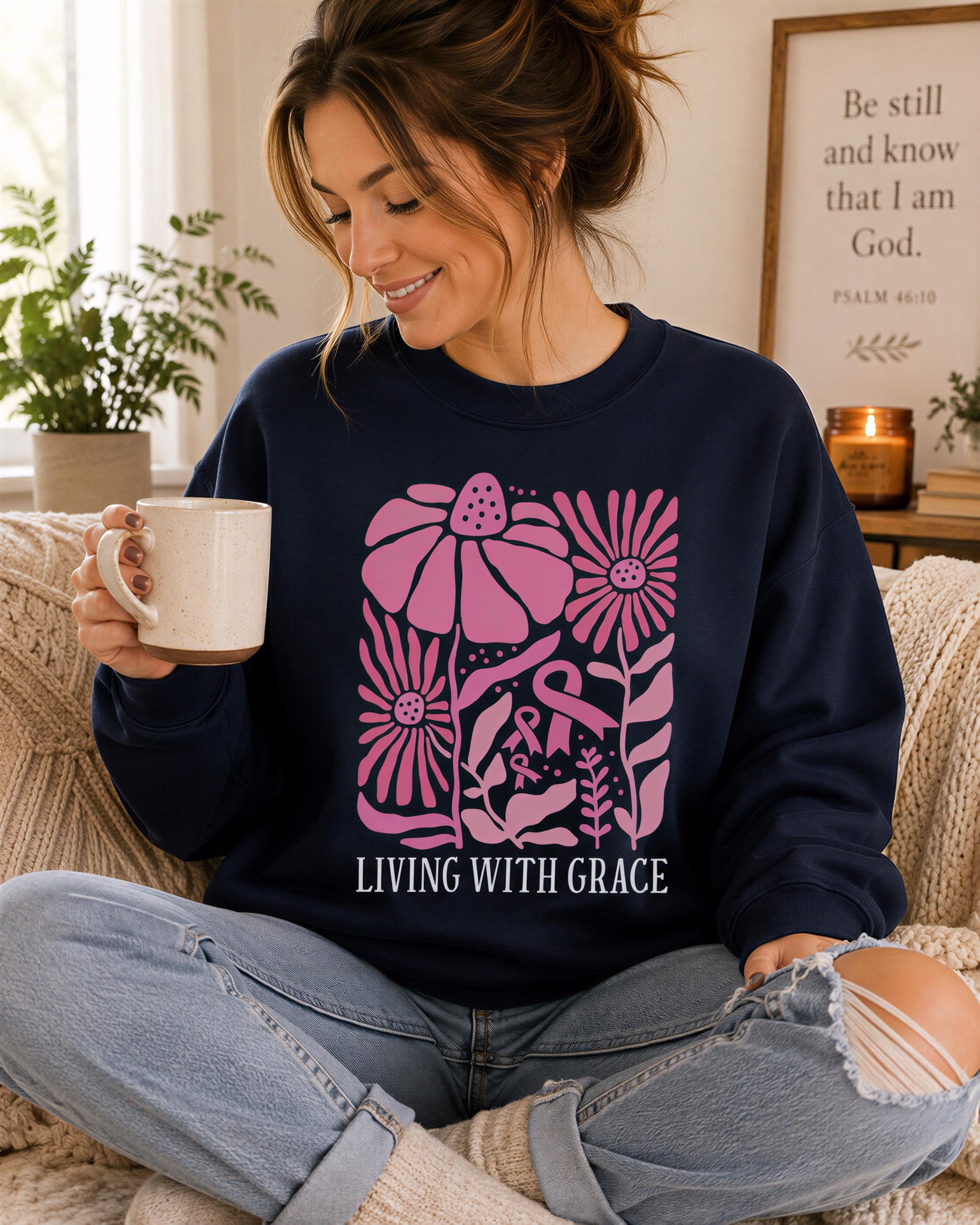 A woman wearing a navy blue sweatshirt with a pink floral design and the text "LIVING WITH GRACE" is sitting on a couch, holding a coffee mug and smiling.