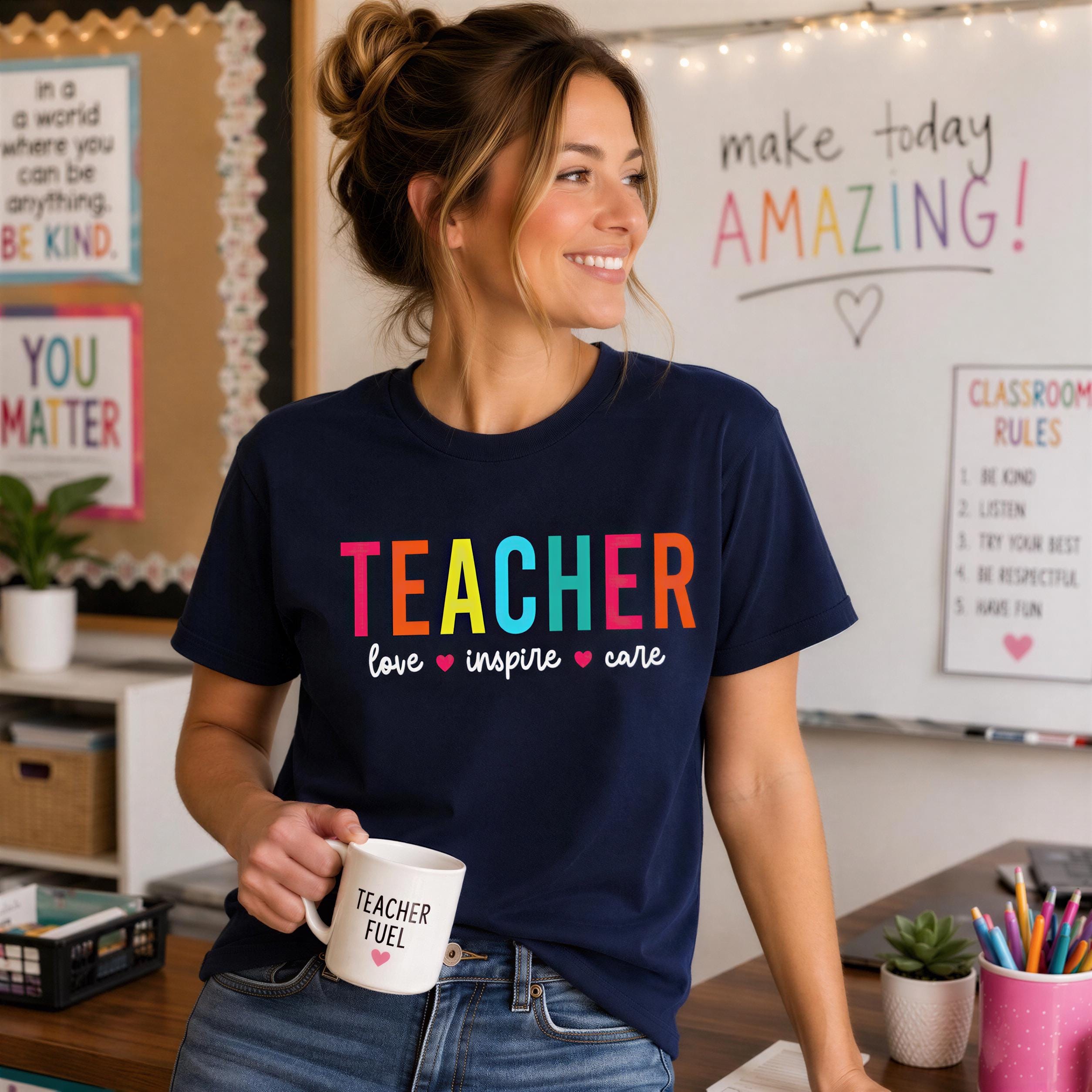 A woman wearing a blue t-shirt with the text "TEACHER love inspire care" is smiling and holding a coffee mug in a classroom setting.