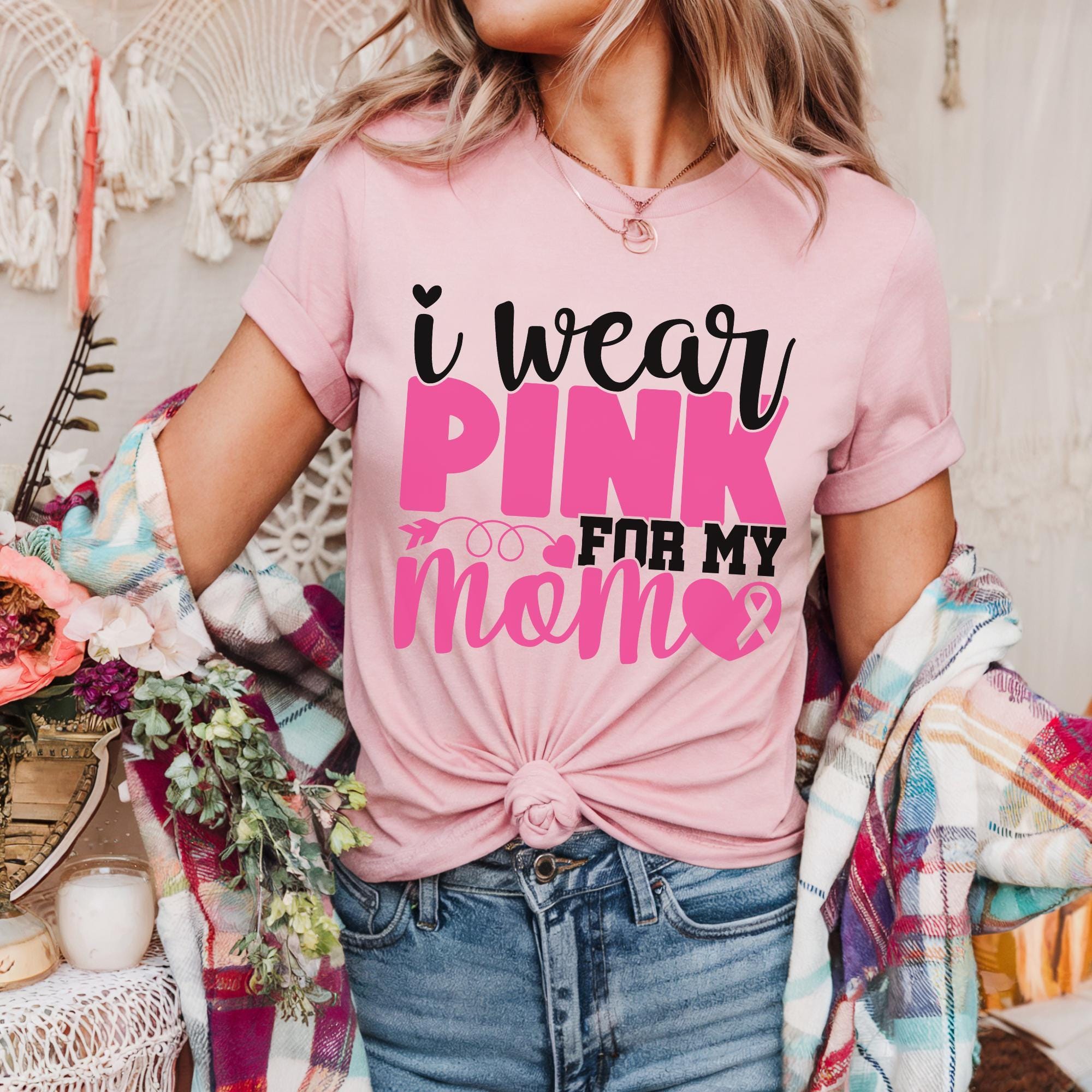 A woman wearing a pink t-shirt with the text "I WEAR PINK FOR MY MOM" printed on it, standing in front of a floral background.