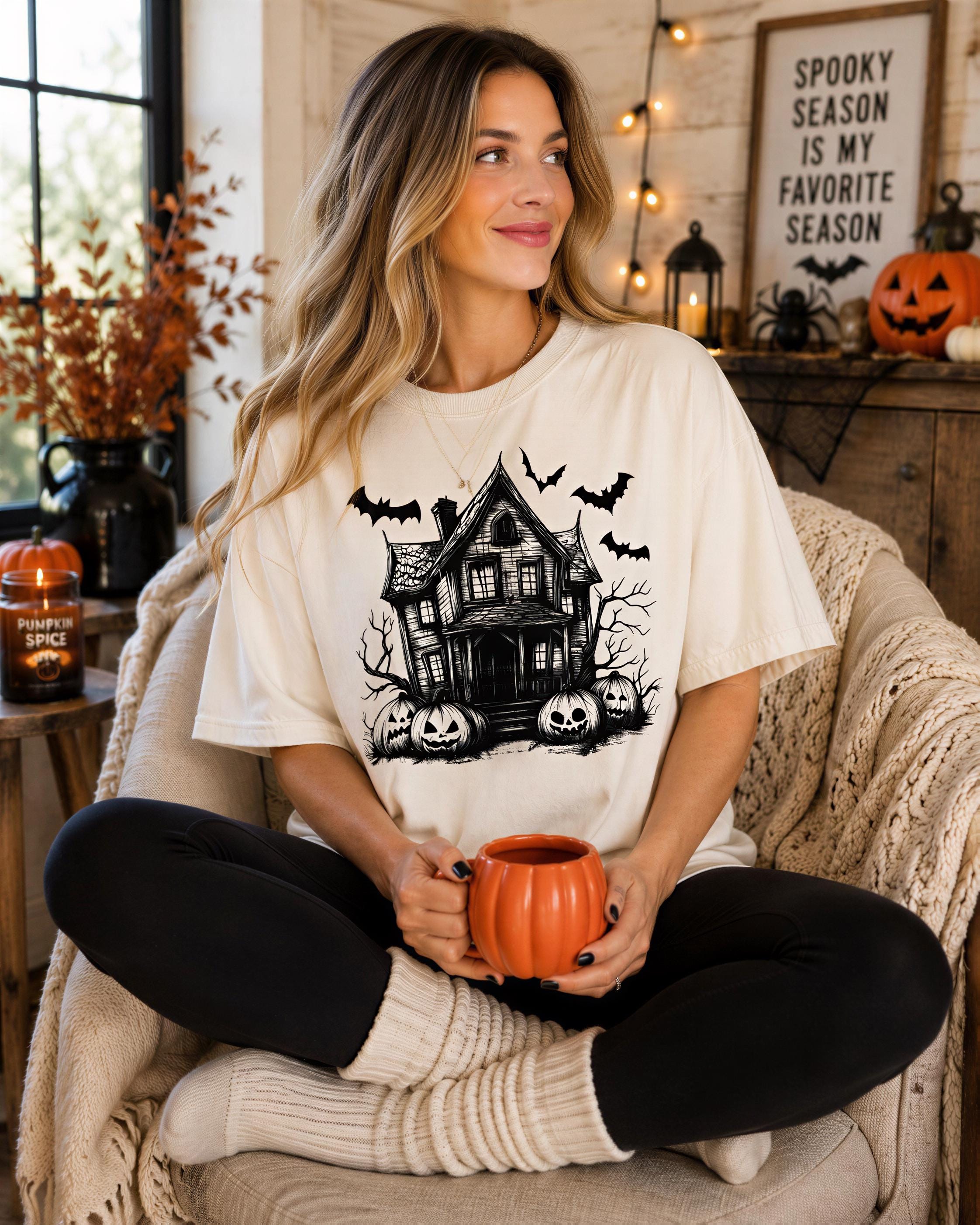 A woman sits in a chair, wearing a white t-shirt with a Halloween-themed graphic, holding a pumpkin-shaped mug and smiling.