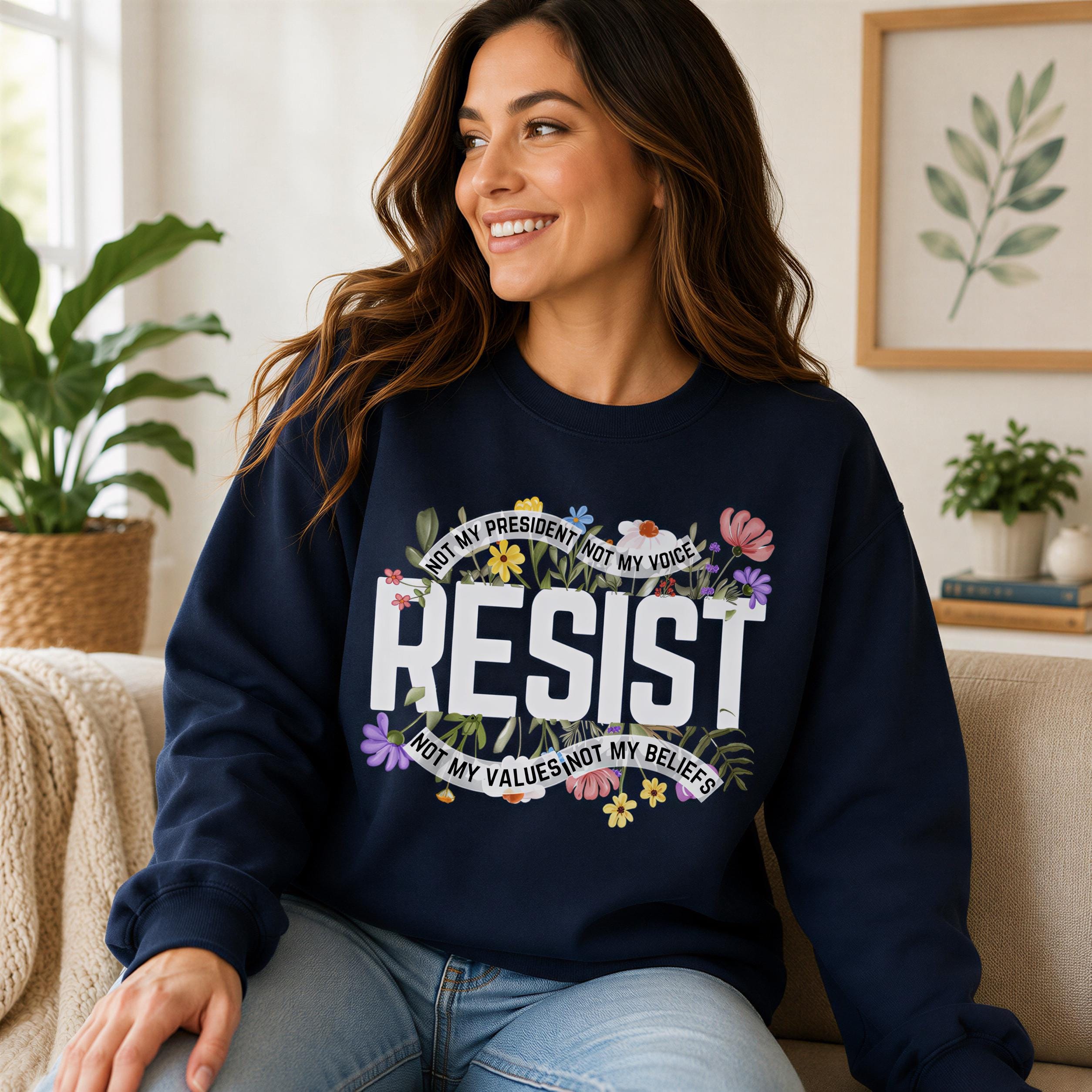A woman wearing a navy blue sweatshirt with a floral design and the text "RESIST NOT MY VOICE NOT MY BELIEFS" is sitting on a couch.