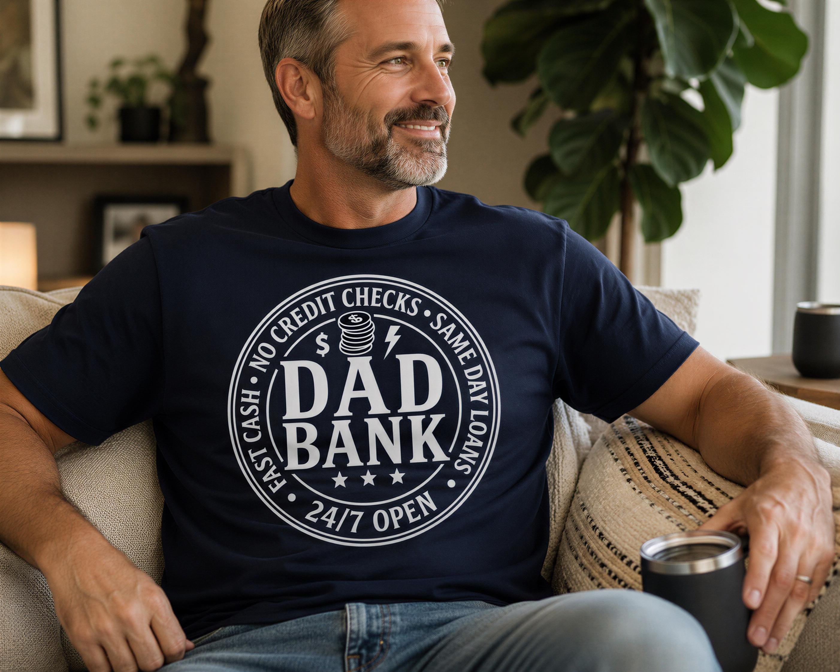 A man wearing a navy blue t-shirt with a humorous text and graphic design is sitting on a couch, smiling and holding a black cup.