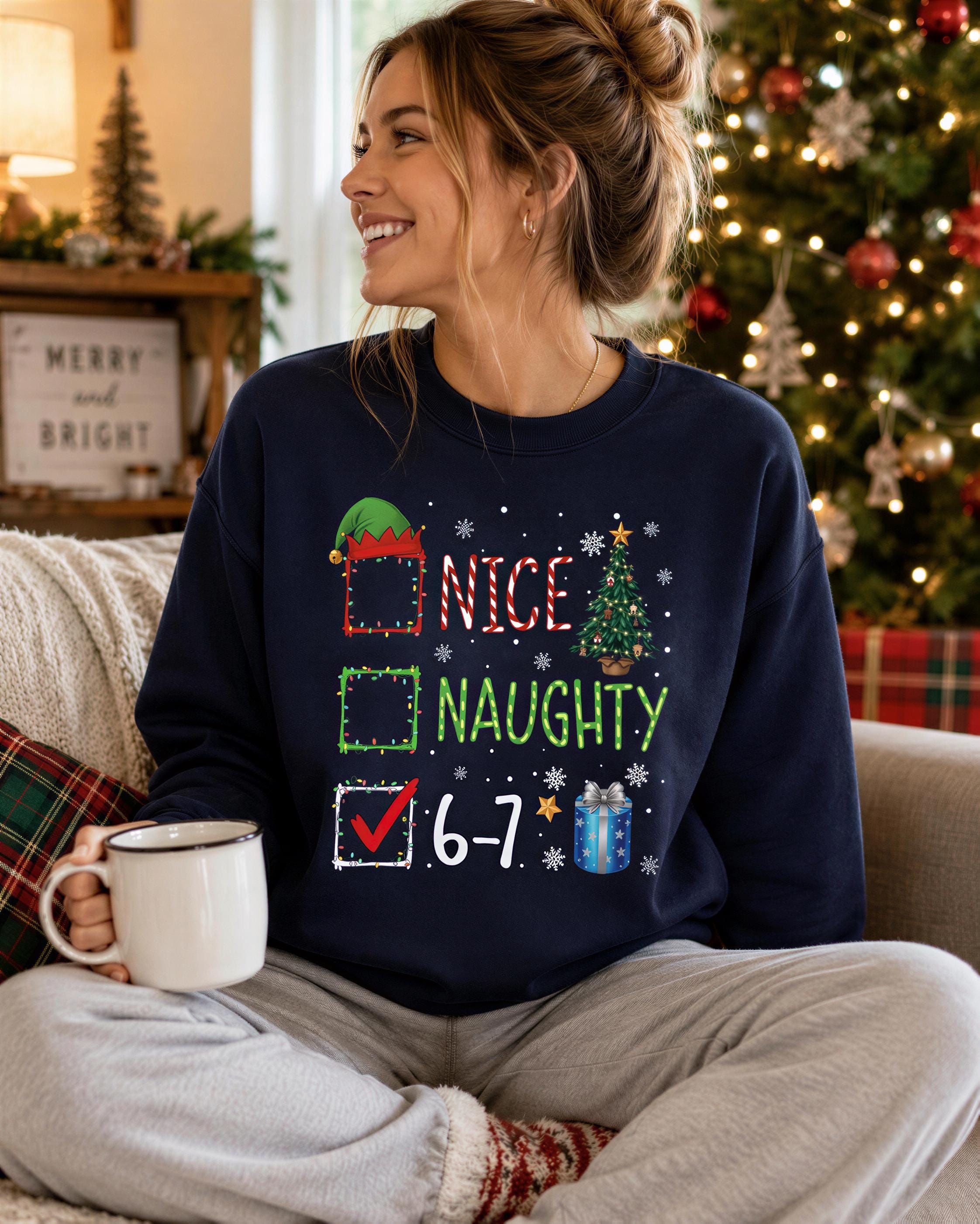 A woman wearing a navy blue sweatshirt with a humorous Christmas-themed design, including a list of Christmas-related words and a checkbox. She is sitting on a couch and holding a coffee mug, with a Christmas tree visible in the background.