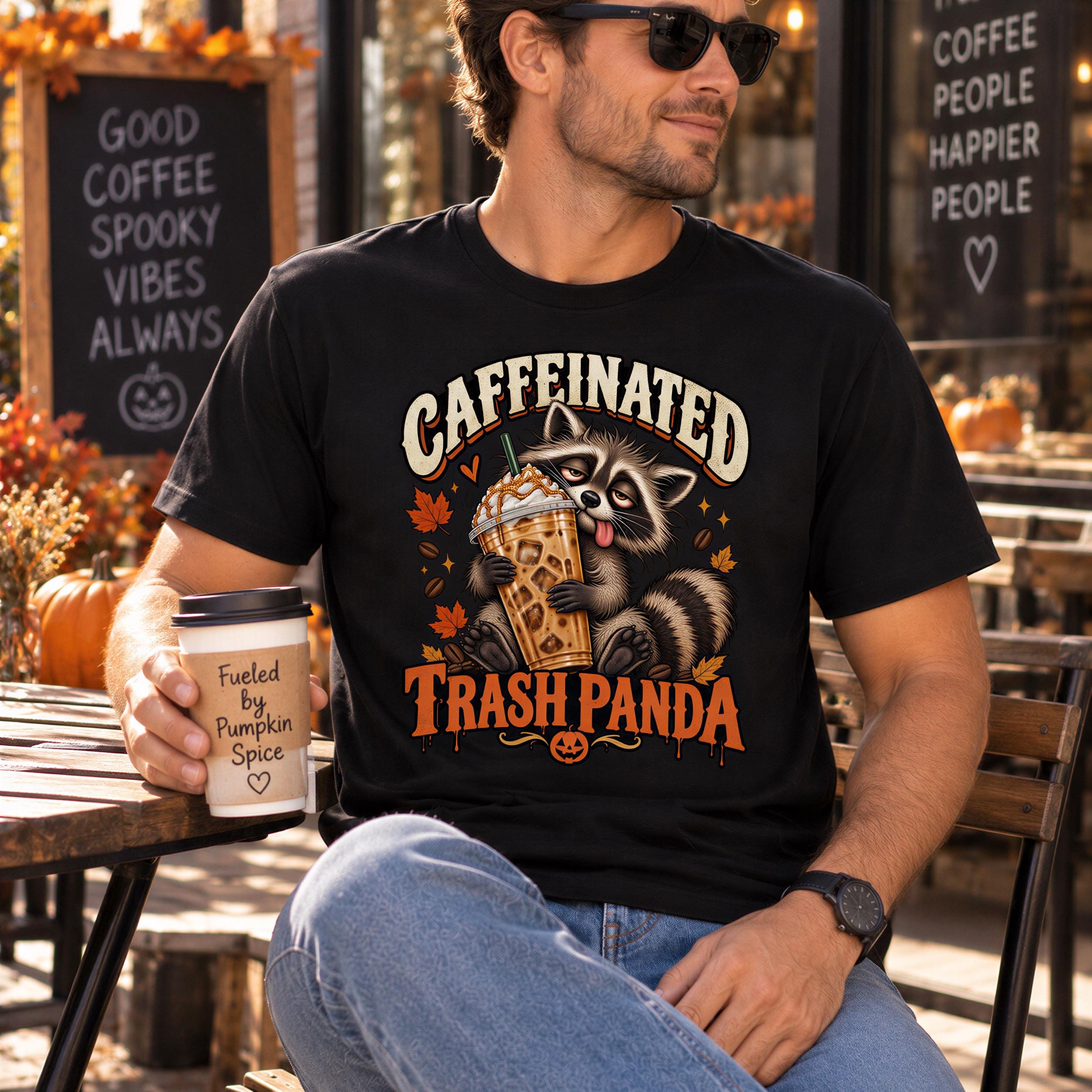 A man wearing a black t-shirt with a graphic of a raccoon holding a coffee drink and the text "Caffeinated Trash Panda" is sitting at a table with a coffee cup in his hand.