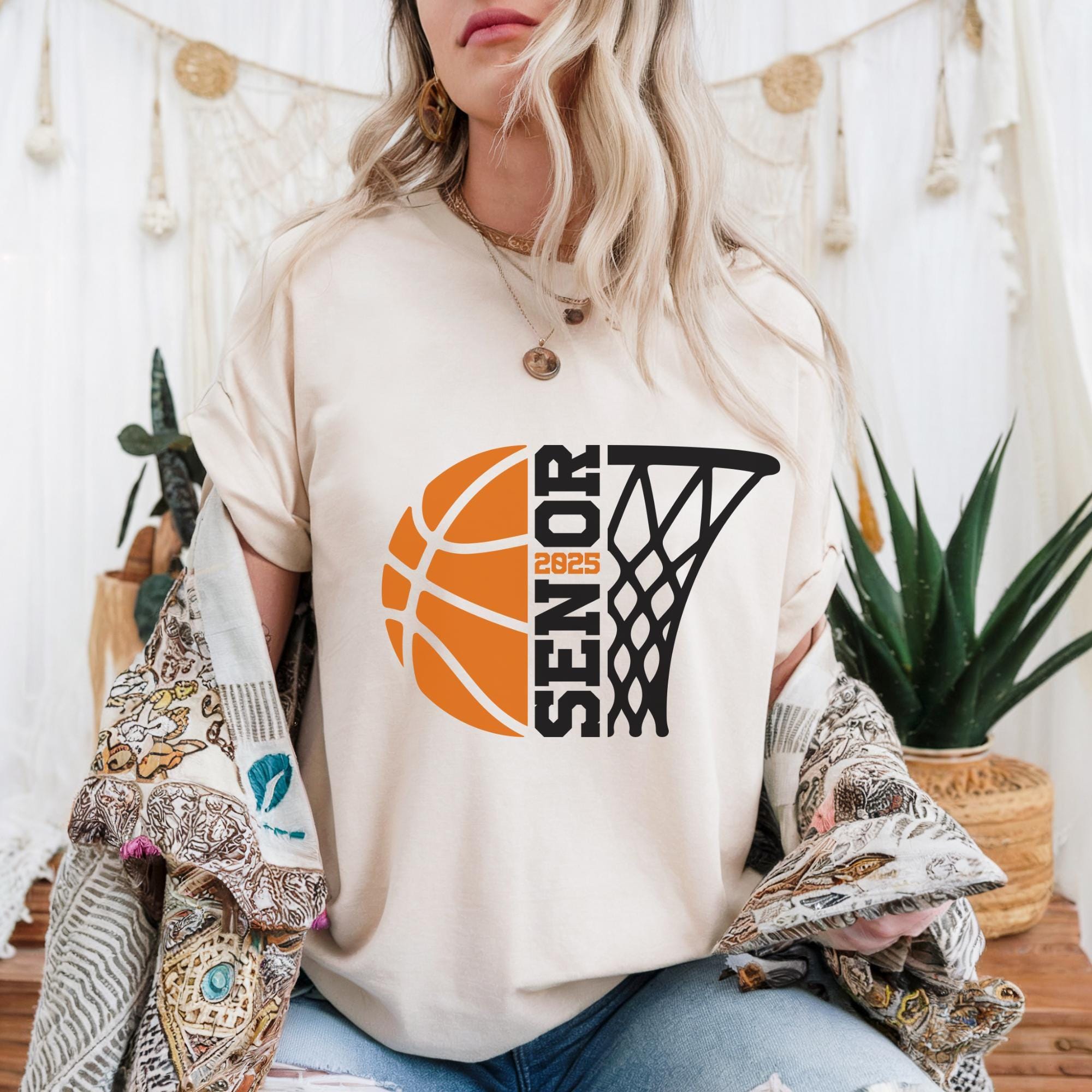 A woman wearing a white t-shirt with a basketball graphic and text, sitting in front of a white curtain with plants and a beaded curtain.