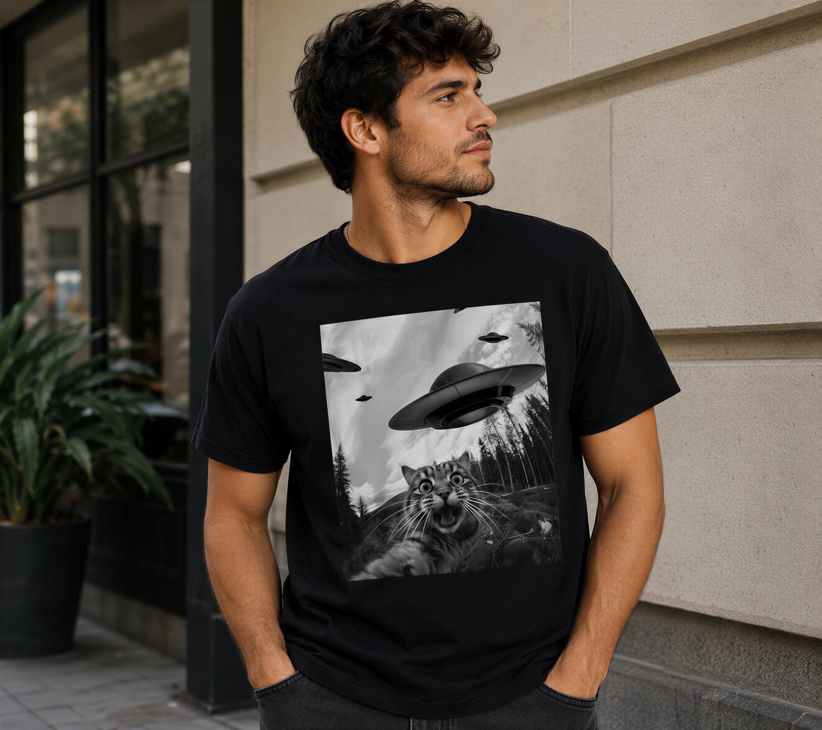 A man wearing a black t-shirt with a graphic of a cat and flying saucers on it, standing in front of a building.