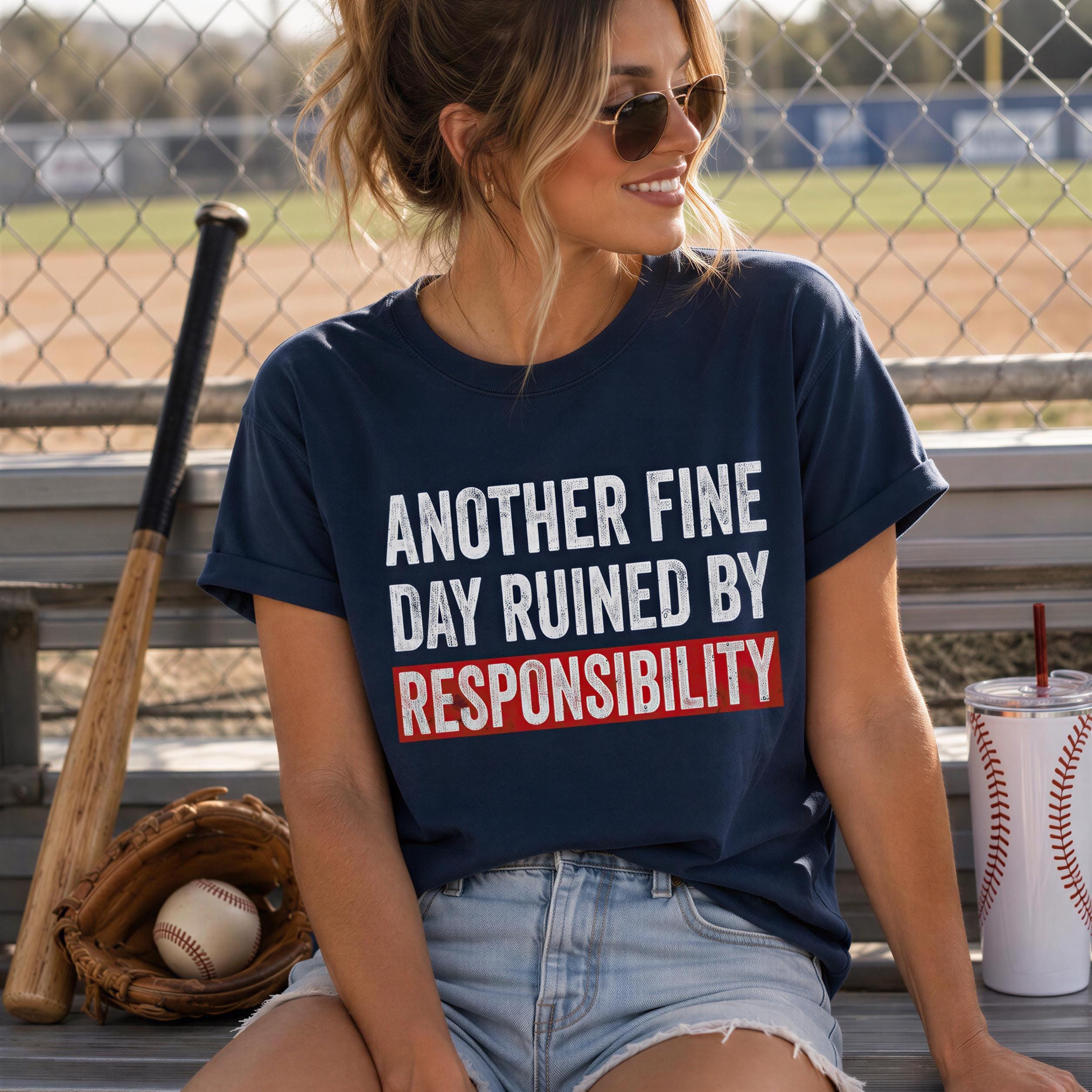 A woman wearing a navy blue t-shirt with the text "Another Fine Day Ruined by Responsibility" sits on a bench with a baseball bat and glove nearby.