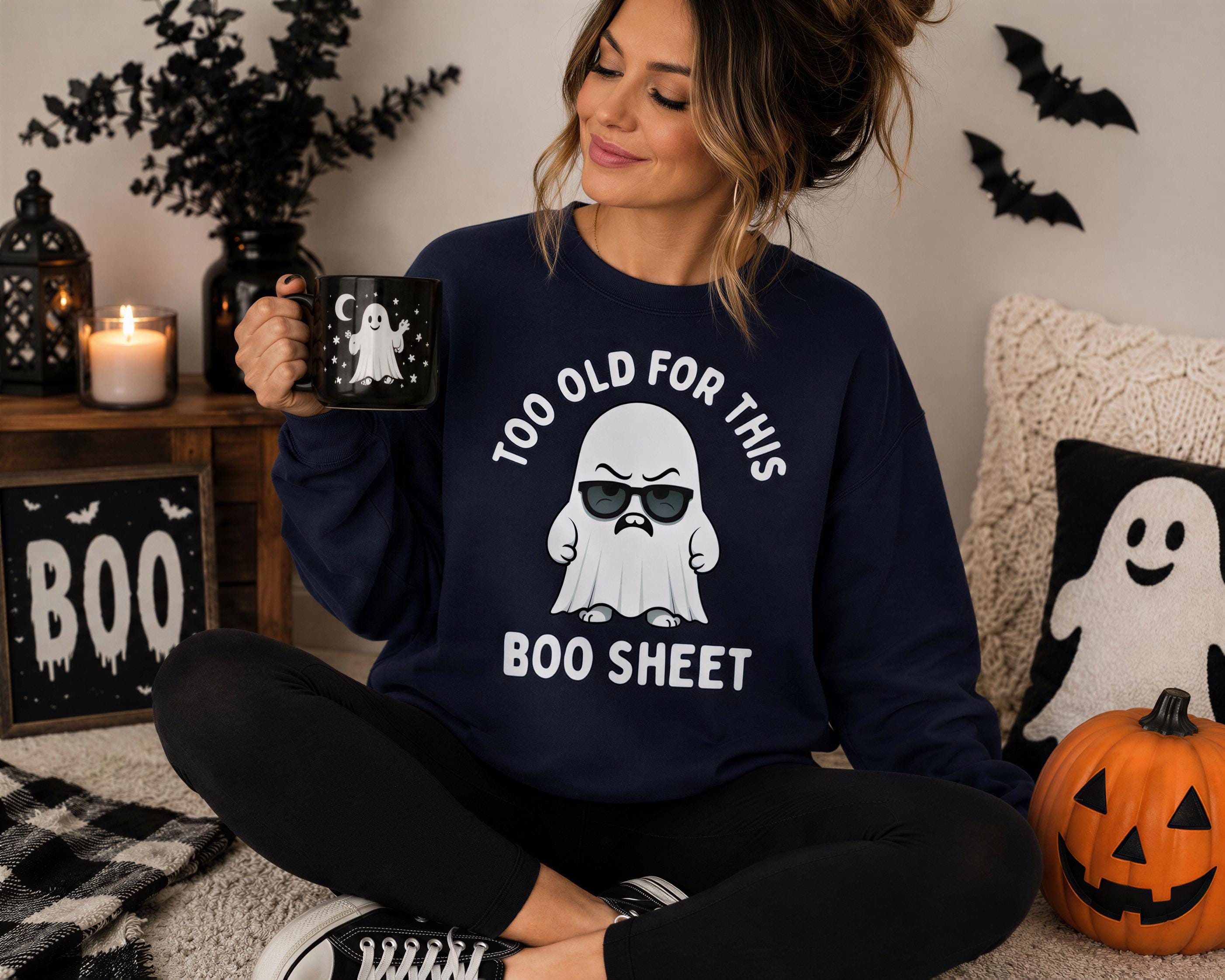 A woman sitting on the floor wearing a navy blue sweatshirt with a graphic of a ghost and the text "Too old for this BOO sheet" is holding a mug with a ghost image. The room has Halloween decorations, including a pumpkin and a ghost-themed pillow.