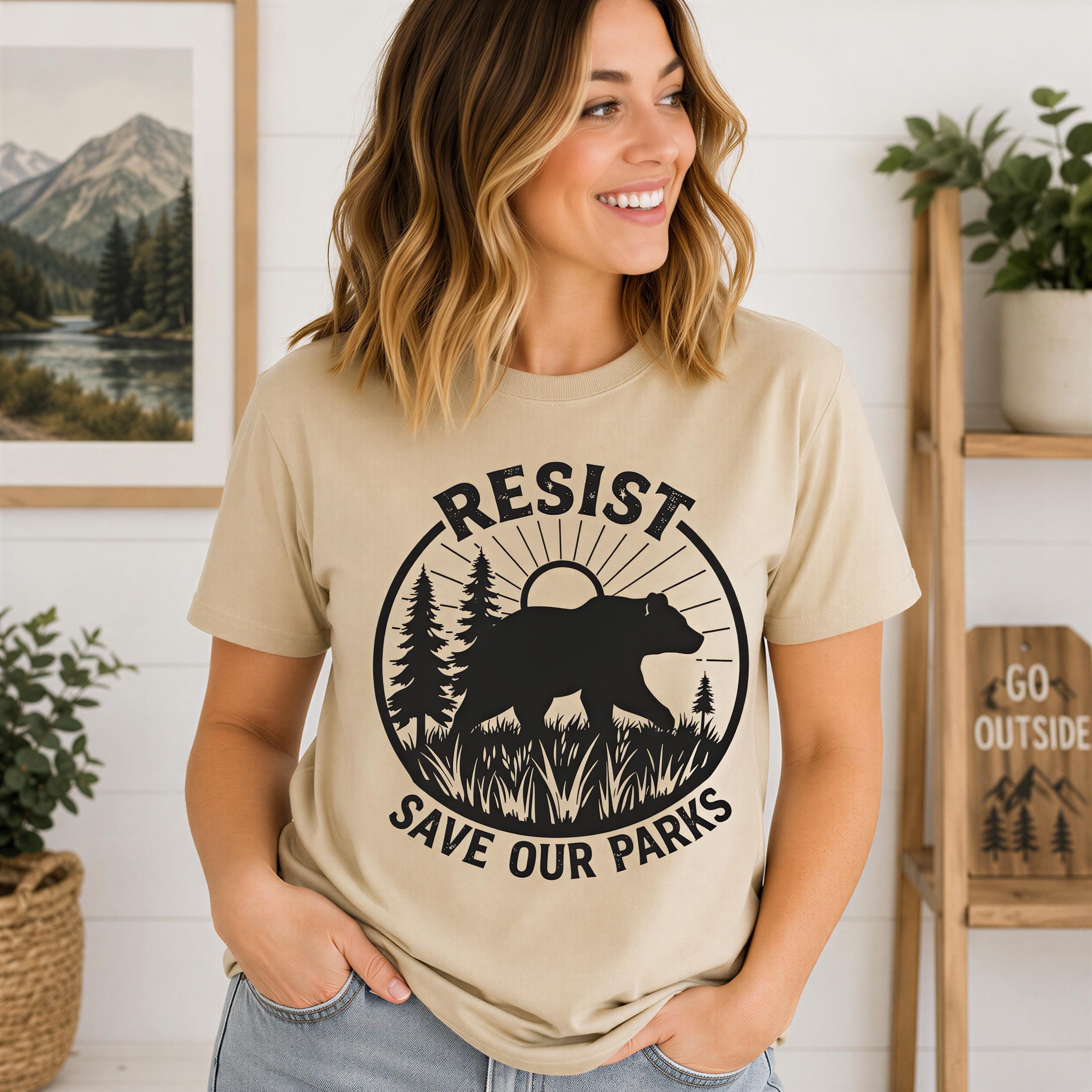 A woman wearing a beige t-shirt with a graphic design of a bear and the text "RESIST SAVE OUR PARKS" is smiling and posing in front of a white wall with framed pictures and plants.
