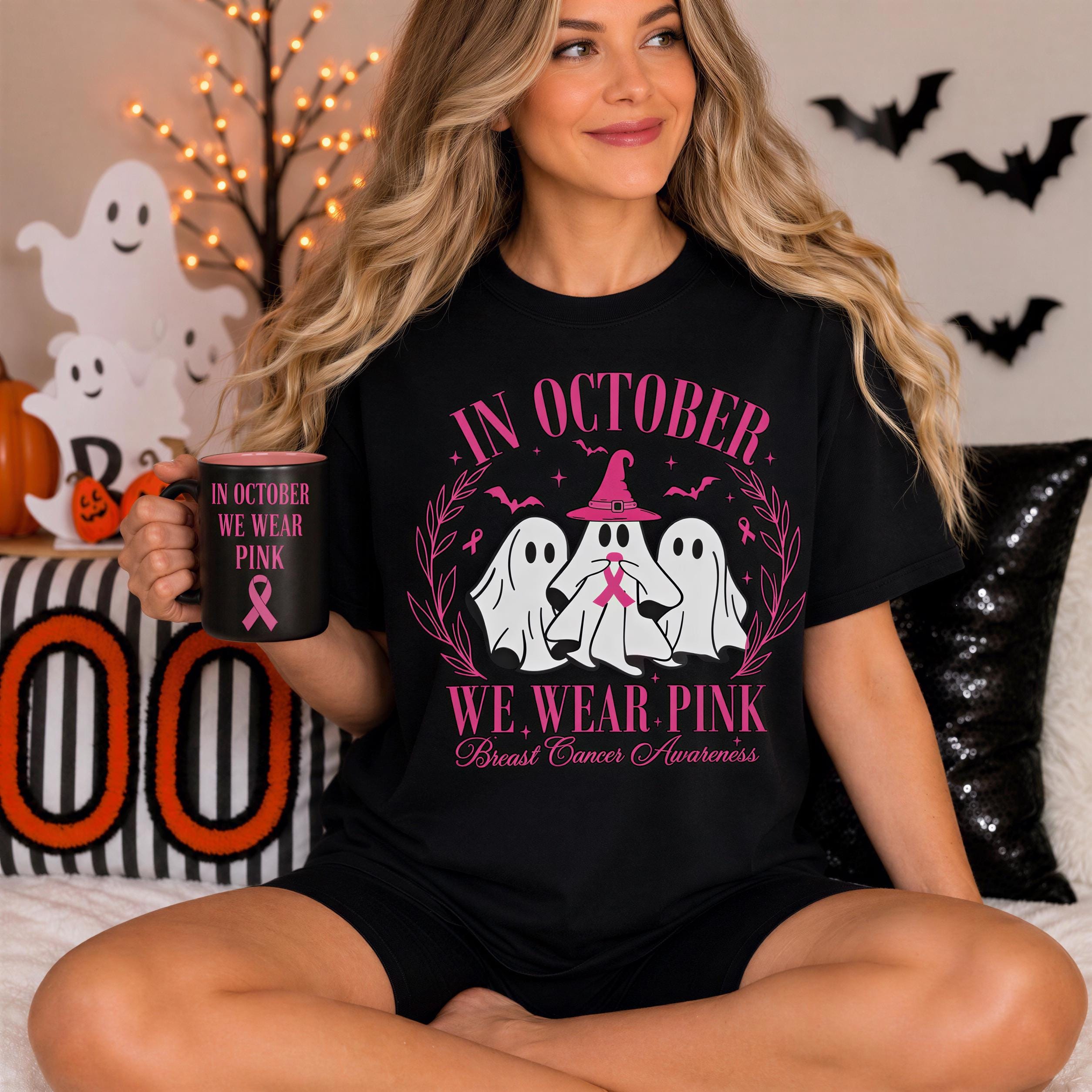 A woman wearing a black t-shirt with a pink design and text, sitting on a bed with Halloween decorations in the background.