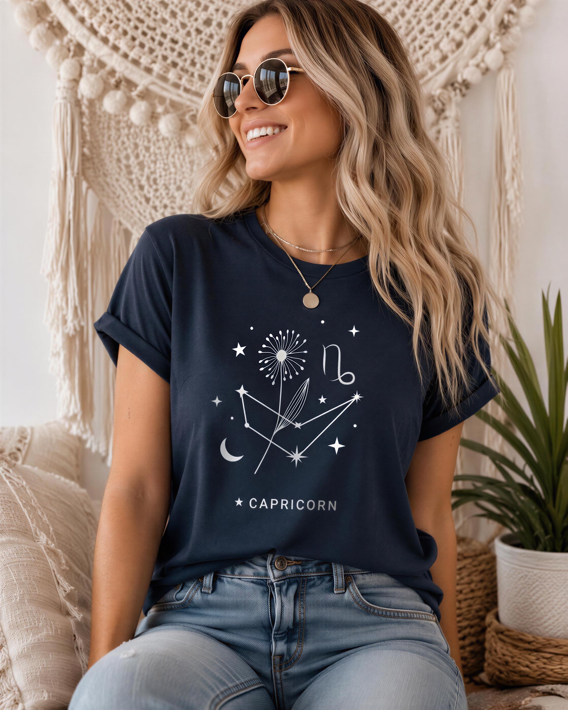 A woman with long blonde hair is sitting on a couch, wearing a navy blue t-shirt with a white graphic design and the word "CAPRICORN" printed on it. She is smiling and looking to the side.