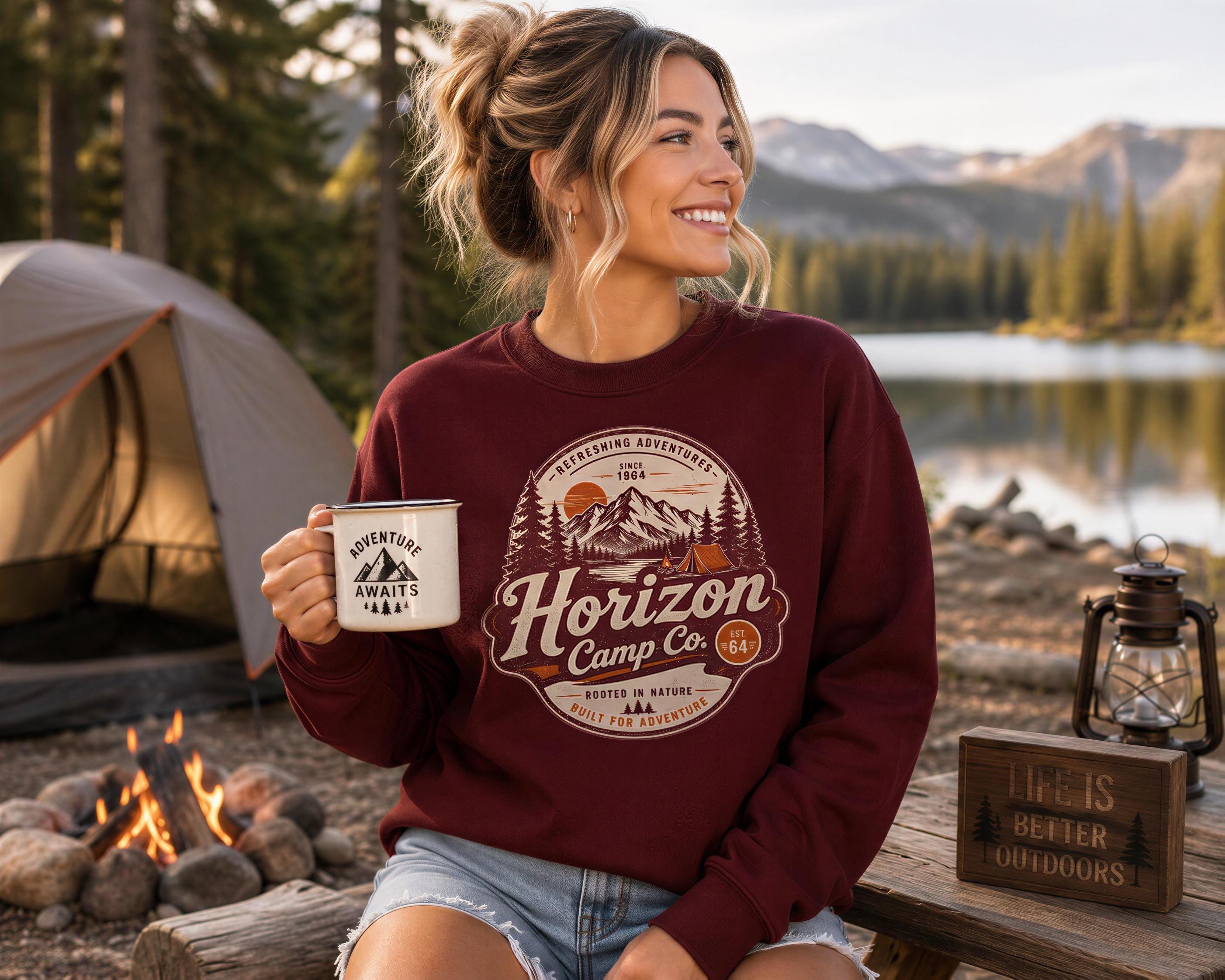 A woman in a maroon sweater sits by a campfire, holding a mug with a mountain and tent design, and smiling at the camera.