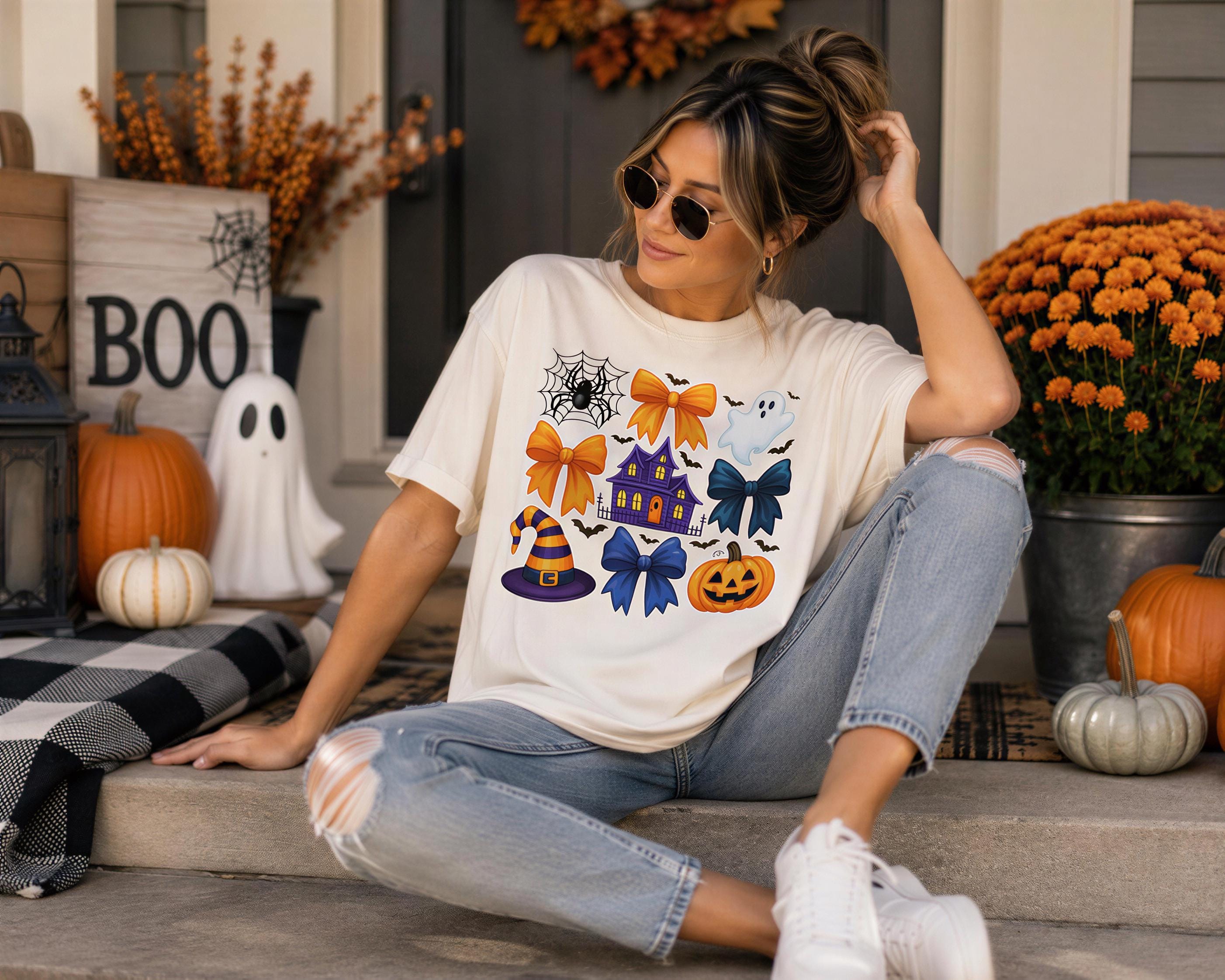 A woman wearing a white t-shirt with Halloween-themed illustrations is sitting on a porch surrounded by Halloween decorations, including pumpkins, a spider web, and a ghost.