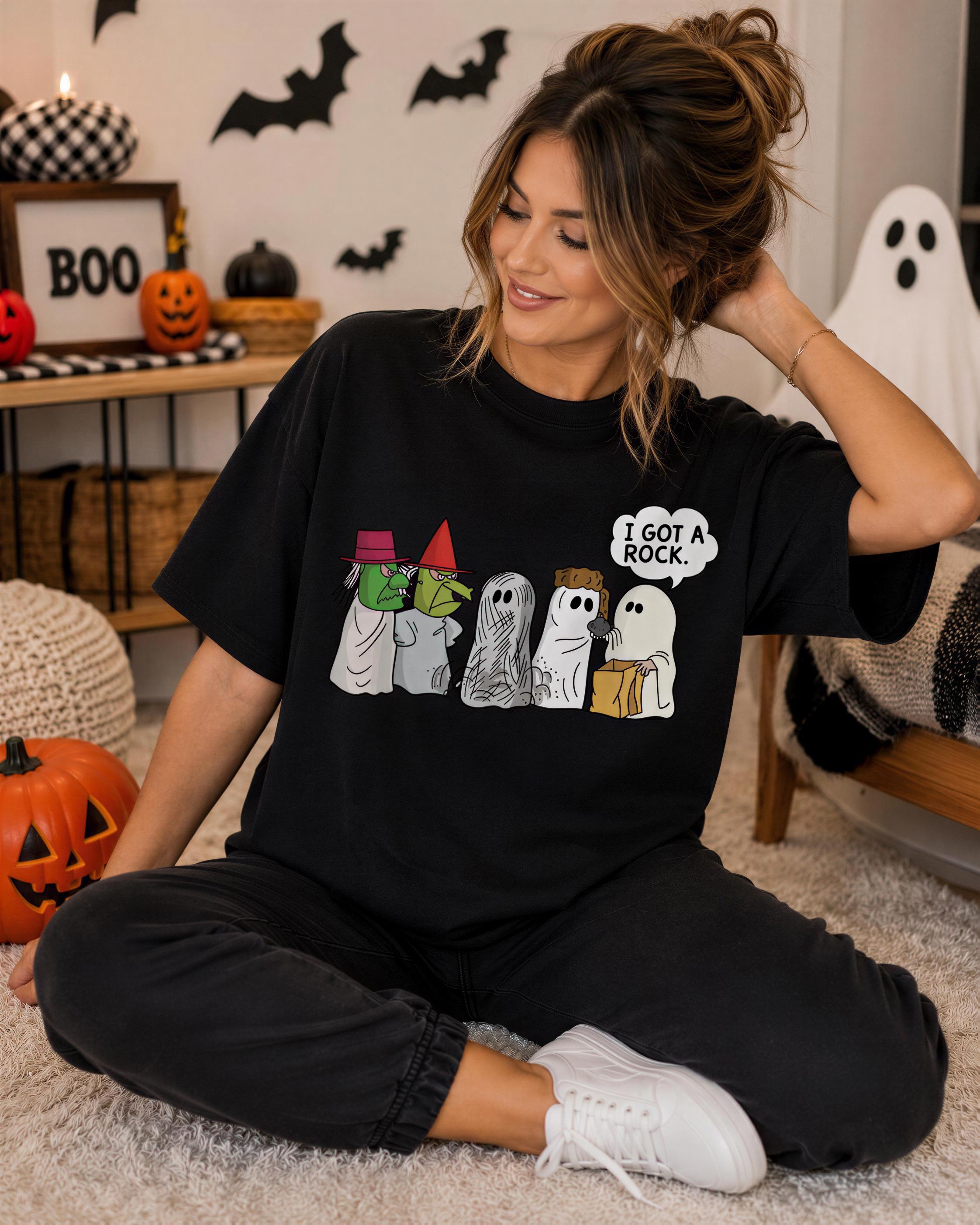 A woman wearing a black t-shirt with a graphic of a group of ghostly figures and text, sitting on the floor in a room decorated for Halloween.