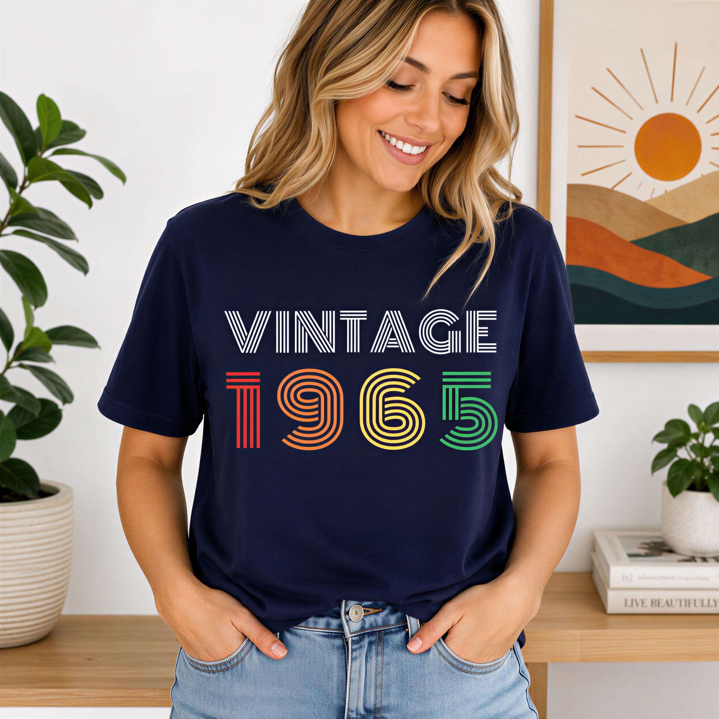 A woman wearing a navy blue t-shirt with the text "VINTAGE 1965" printed on it, smiling and posing in front of a wall with a framed picture.