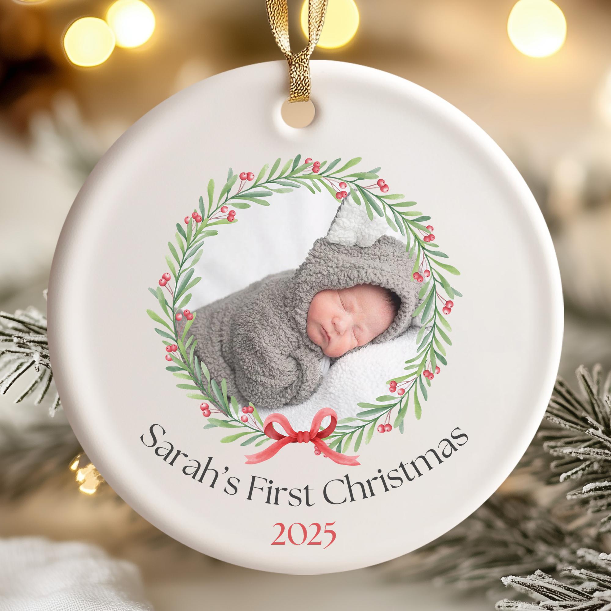 A white ornament with a photo of a sleeping baby and a wreath of greenery and red berries. The text on the ornament reads "Sarah's First Christmas 2025".
