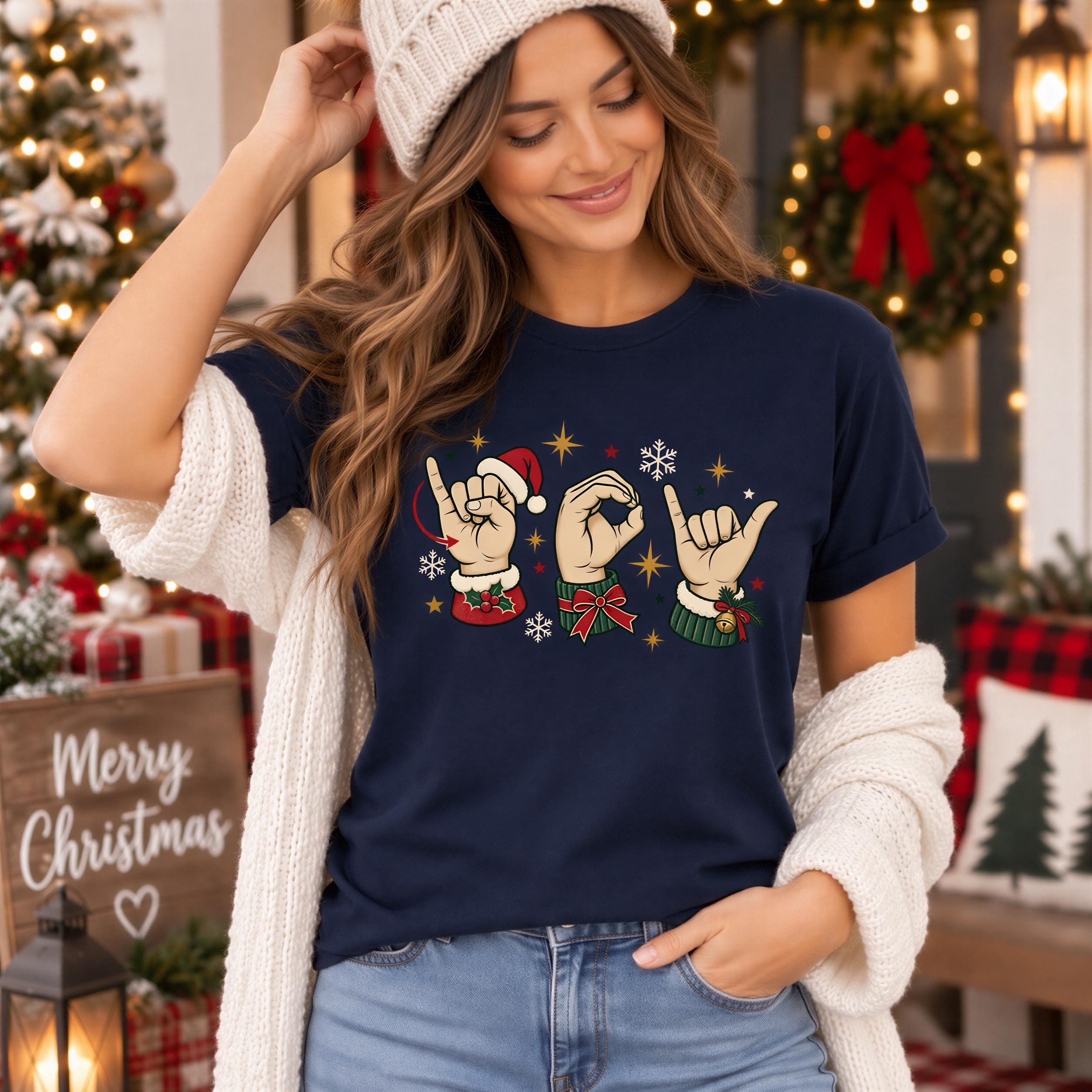 A woman wearing a blue t-shirt with a graphic of a hand making a sign and snowflakes, standing in front of a Christmas tree and wreath.
